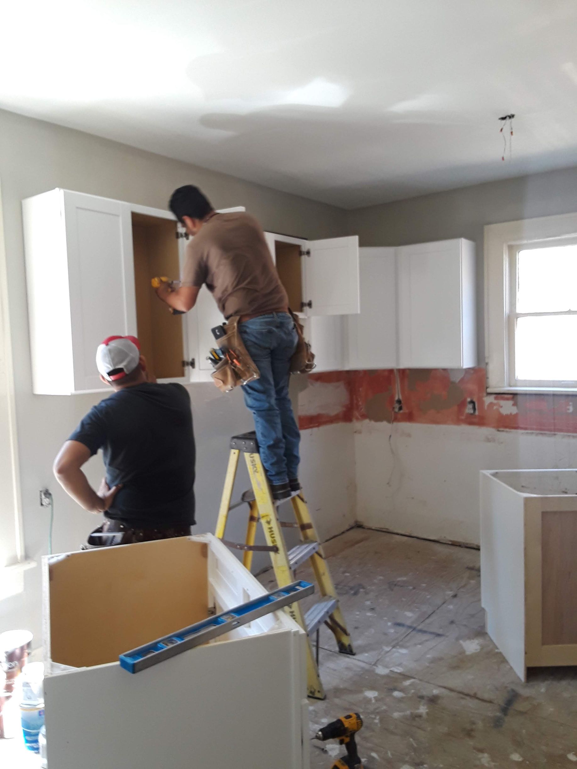 Two people installing white kitchen cabinets in a room under construction; one on a ladder.