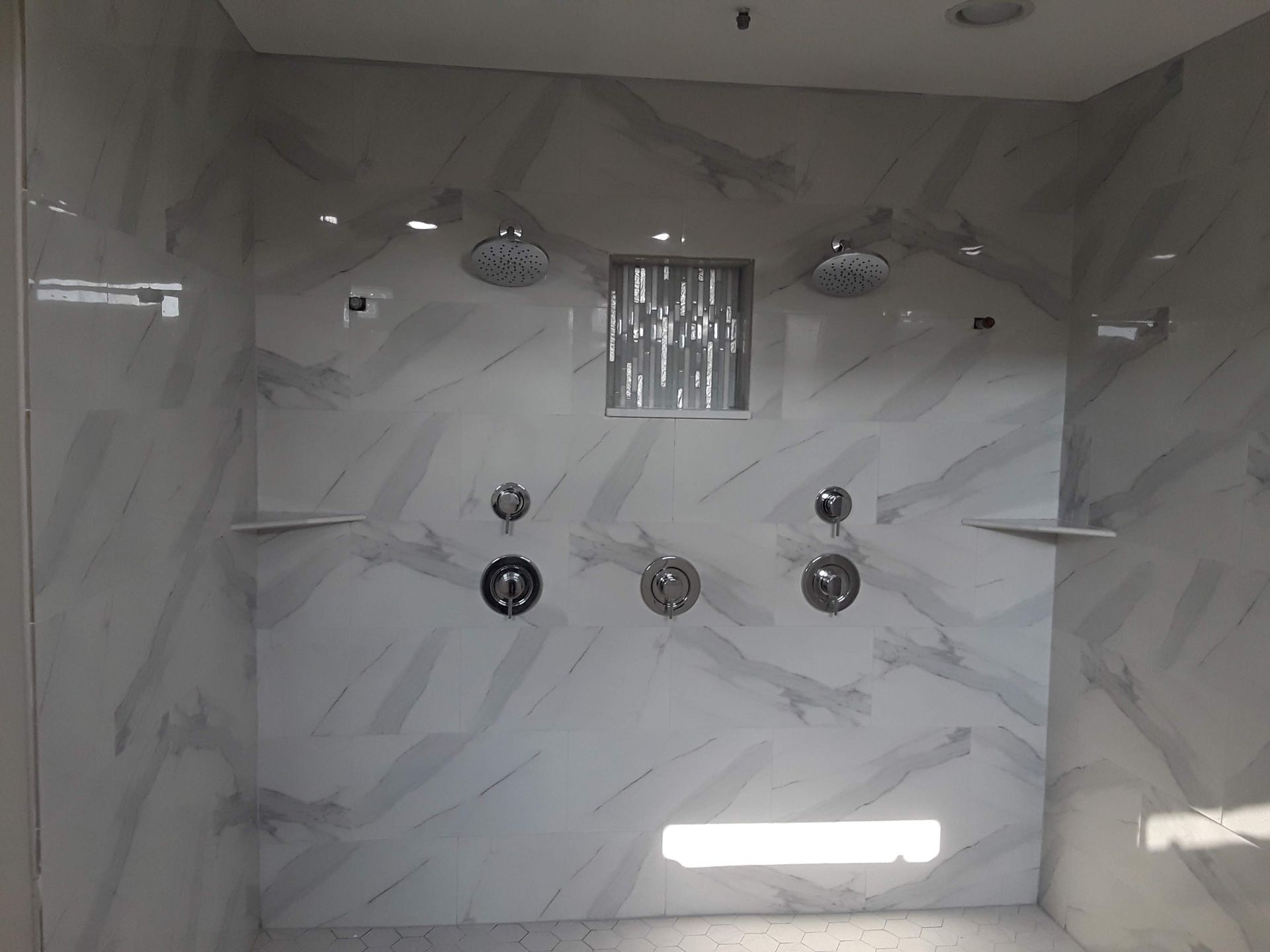 White marble-tiled shower with two shower heads, three faucet controls, a built-in shelf, and recessed lighting.