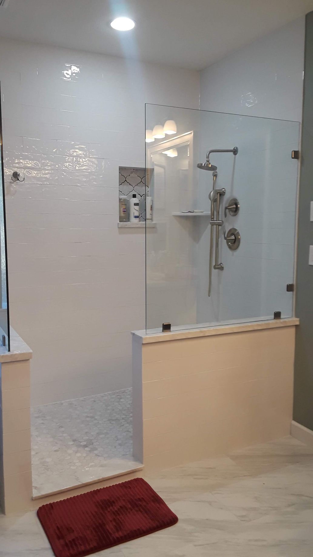 Modern white tiled shower with glass doors, a built-in shelf, and a red bath mat.
