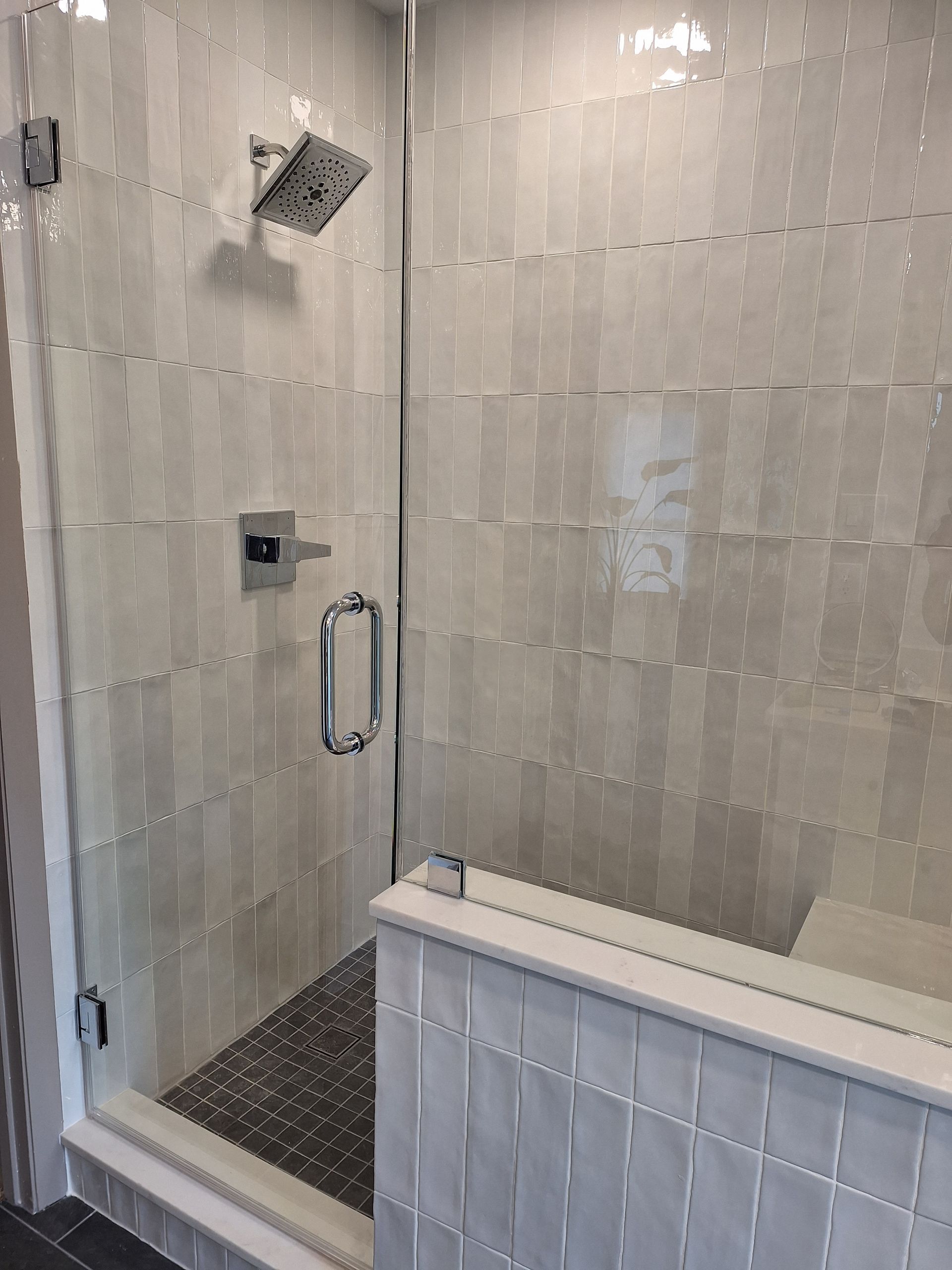 Modern shower with glass door and white tiled walls. Chrome fixtures, small bench, and dark floor.