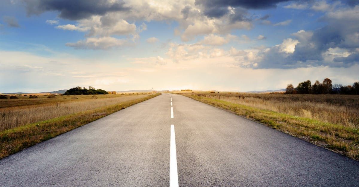 An empty road going through a field with a cloudy sky in the background.