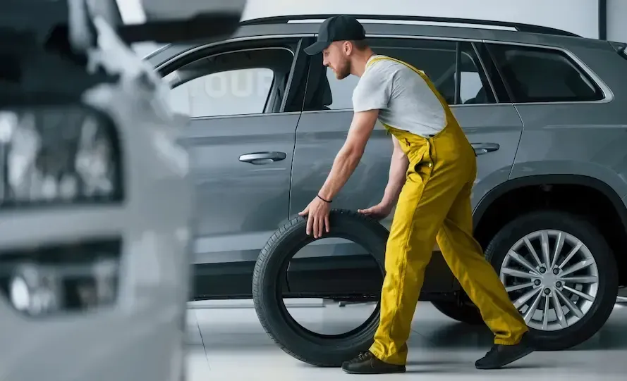 Mecânico de macacão amarelo empurrando um pneu em direção a um SUV cinza em uma garagem.