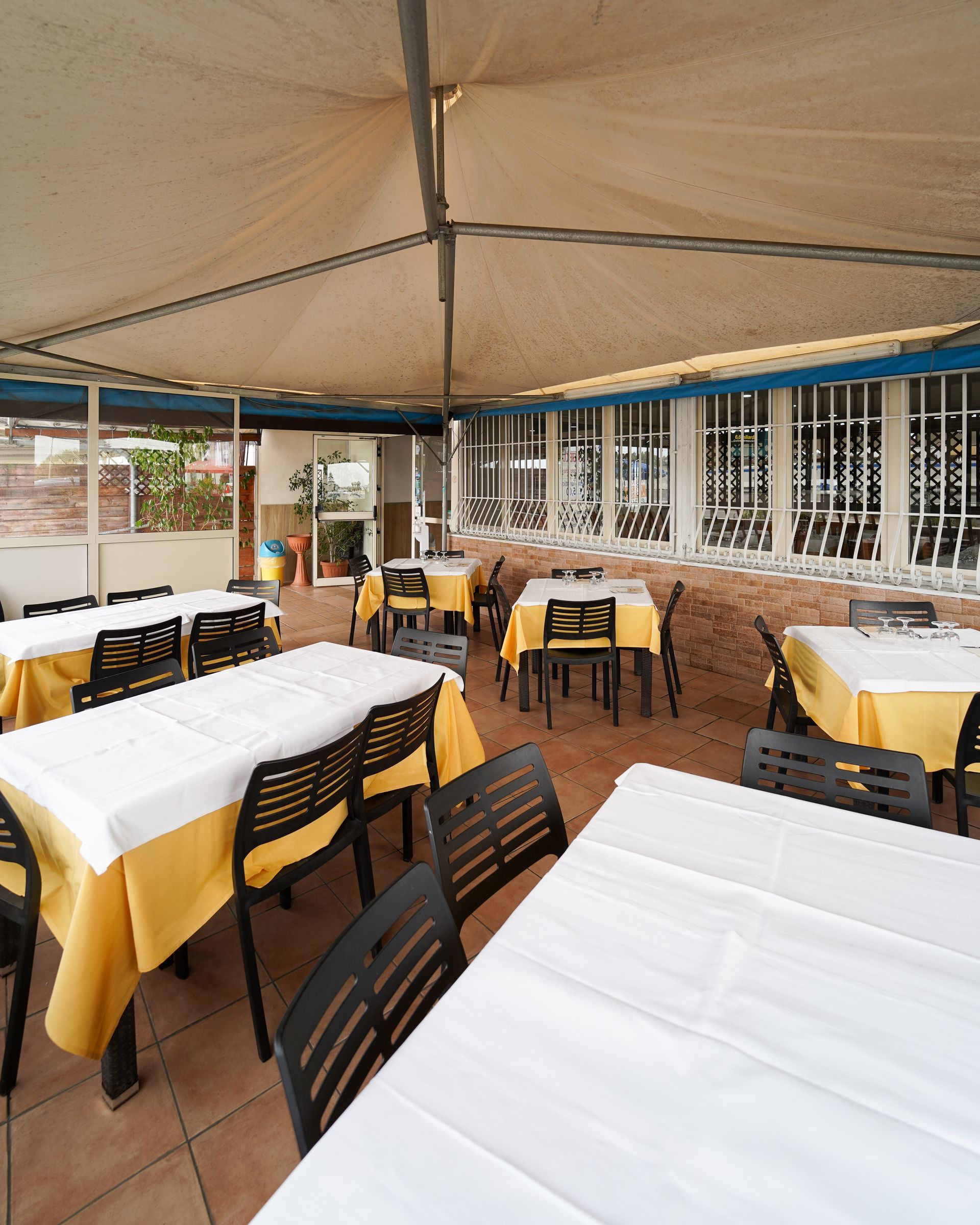 Dehor di un ristorante all'aperto con tovaglie bianche, coprisedie gialli e tavoli sotto una tettoia beige.
