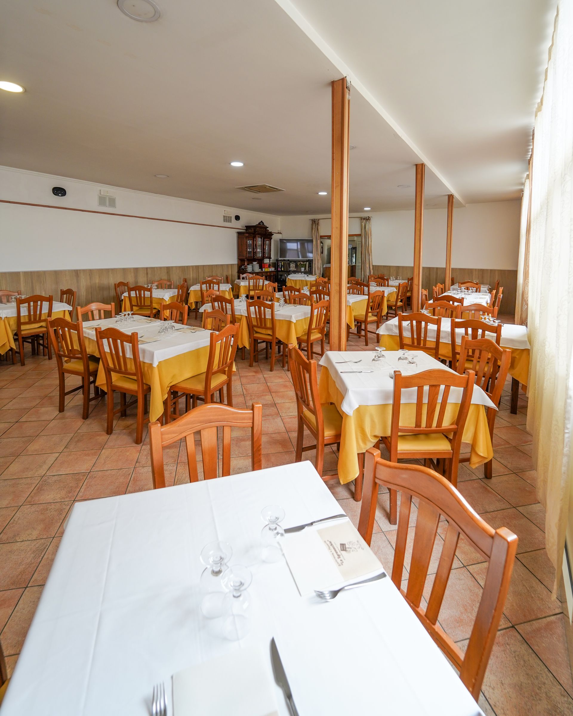 Sala da pranzo vuota di un ristorante con sedie e tavoli in legno apparecchiati con tovaglie bianche.