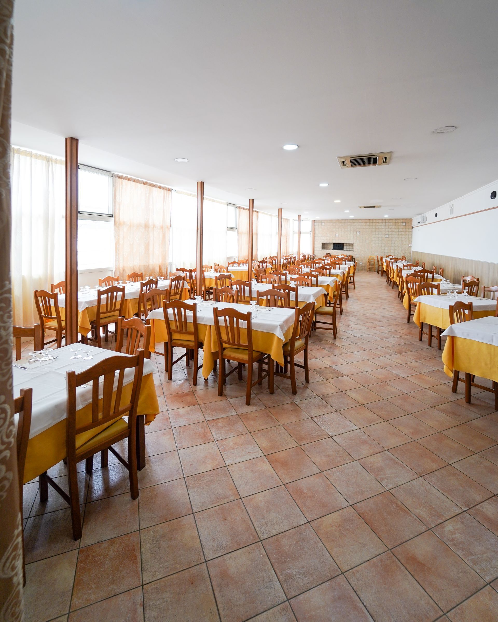 Sala da pranzo vuota con file di sedie e tavoli di legno, pavimento piastrellato e grandi finestre con tende trasparenti.