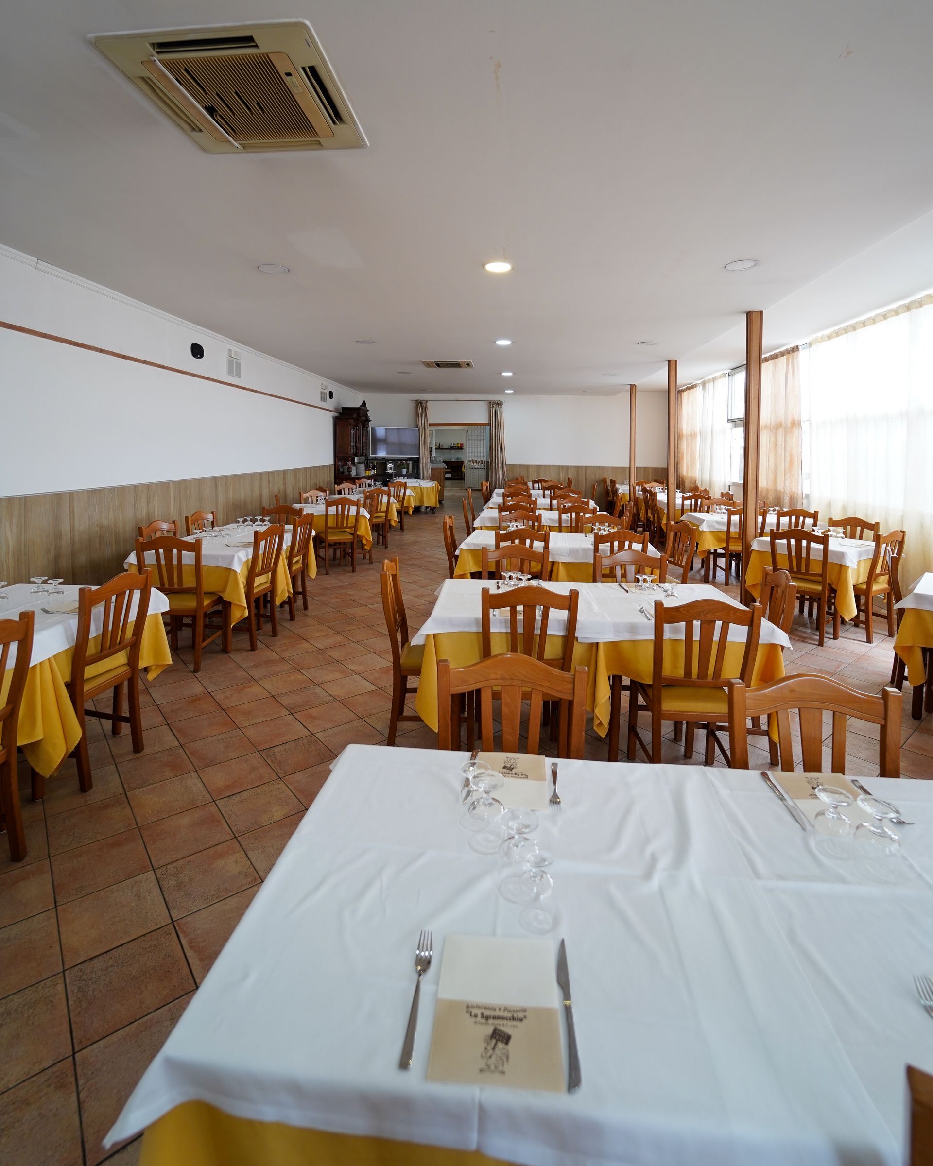 Sala da pranzo vuota di un ristorante con sedie di legno, tovaglie bianche e finestre luminose sul lato destro.