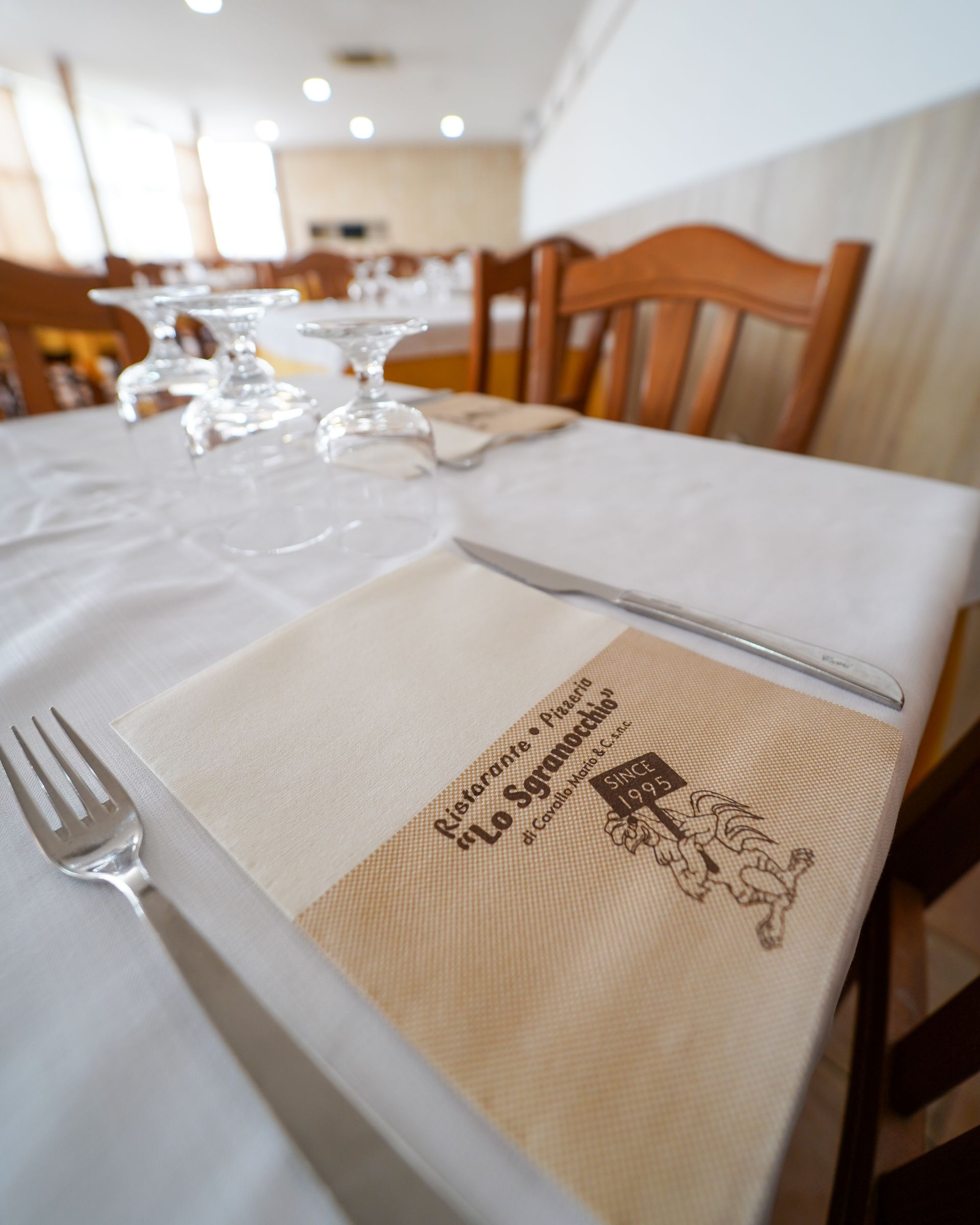 In una luminosa sala da pranzo di un ristorante, apparecchiate la tavola con una tovaglia bianca, un tovagliolo piegato e dei bicchieri.