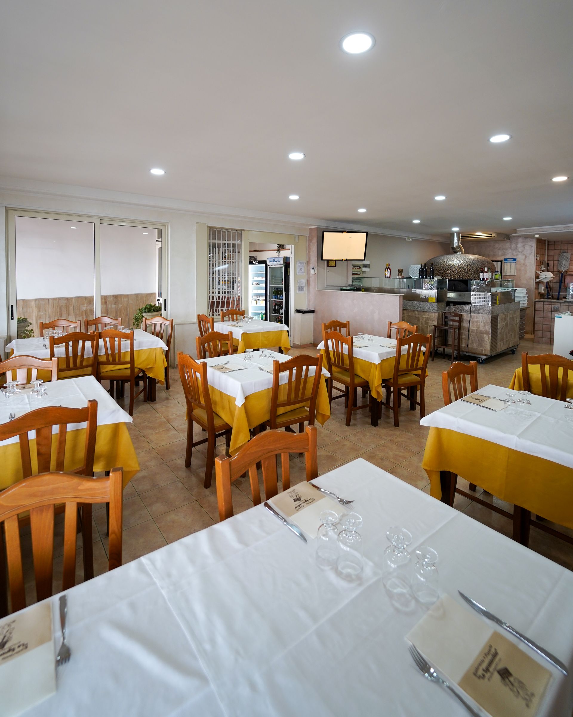 Luminosa sala da pranzo del ristorante con tavoli in legno, sedie gialle e tovaglie bianche illuminate da faretti a incasso.