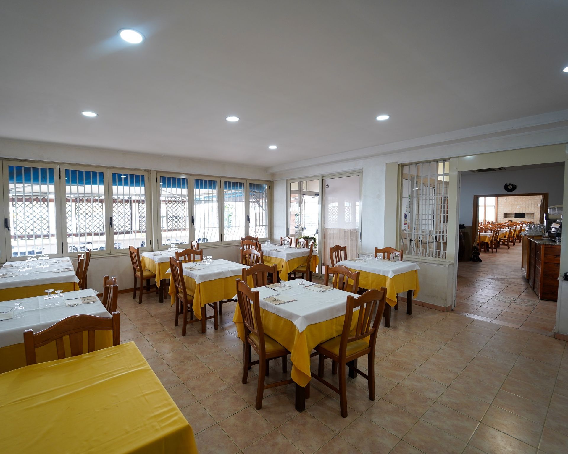 Luminosa sala da pranzo con tovaglie gialle, sedie in legno e ampie finestre lungo la parete sinistra.