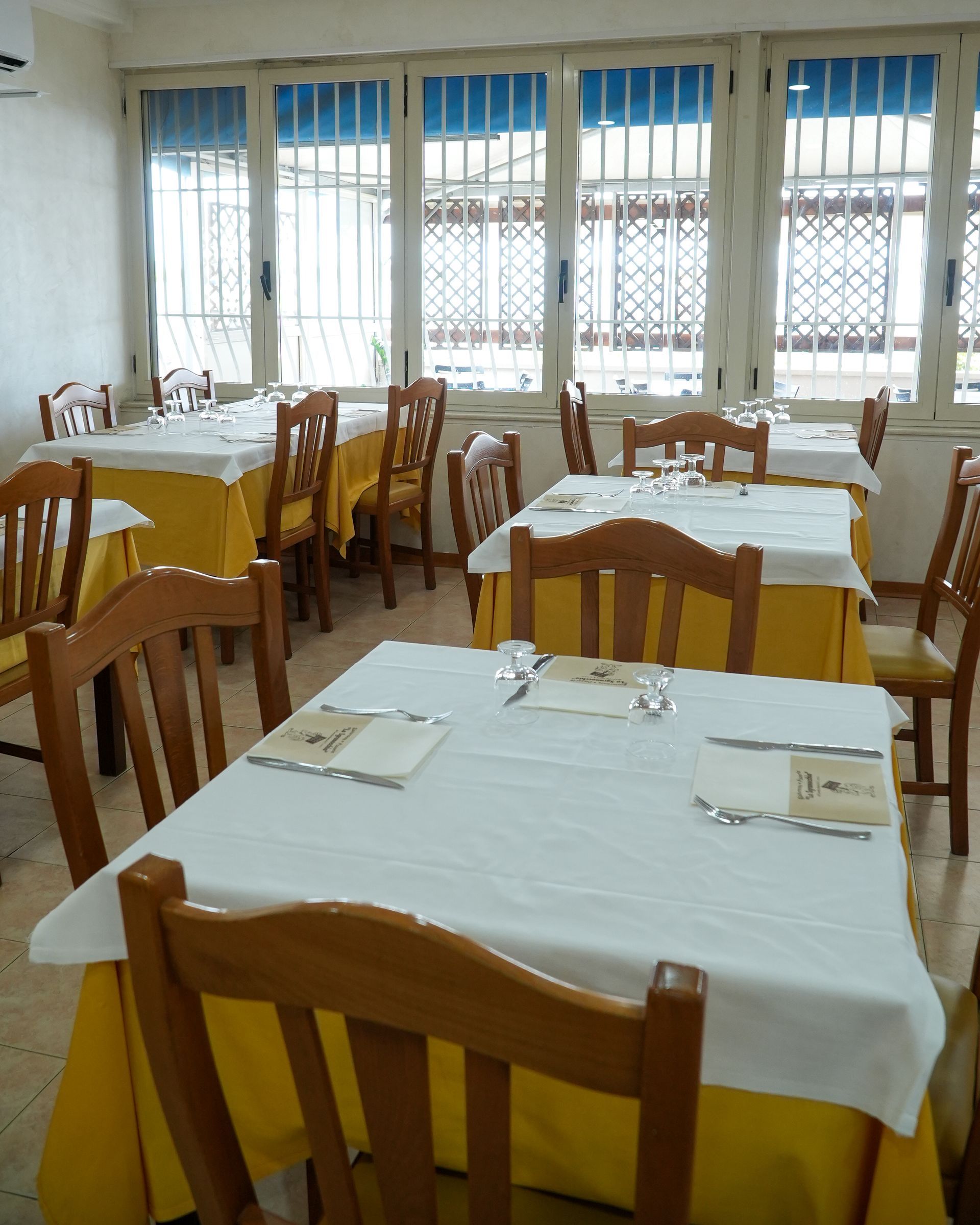 Sala da pranzo con sedie in legno, tovaglie bianche e ampie finestre con tende verticali.