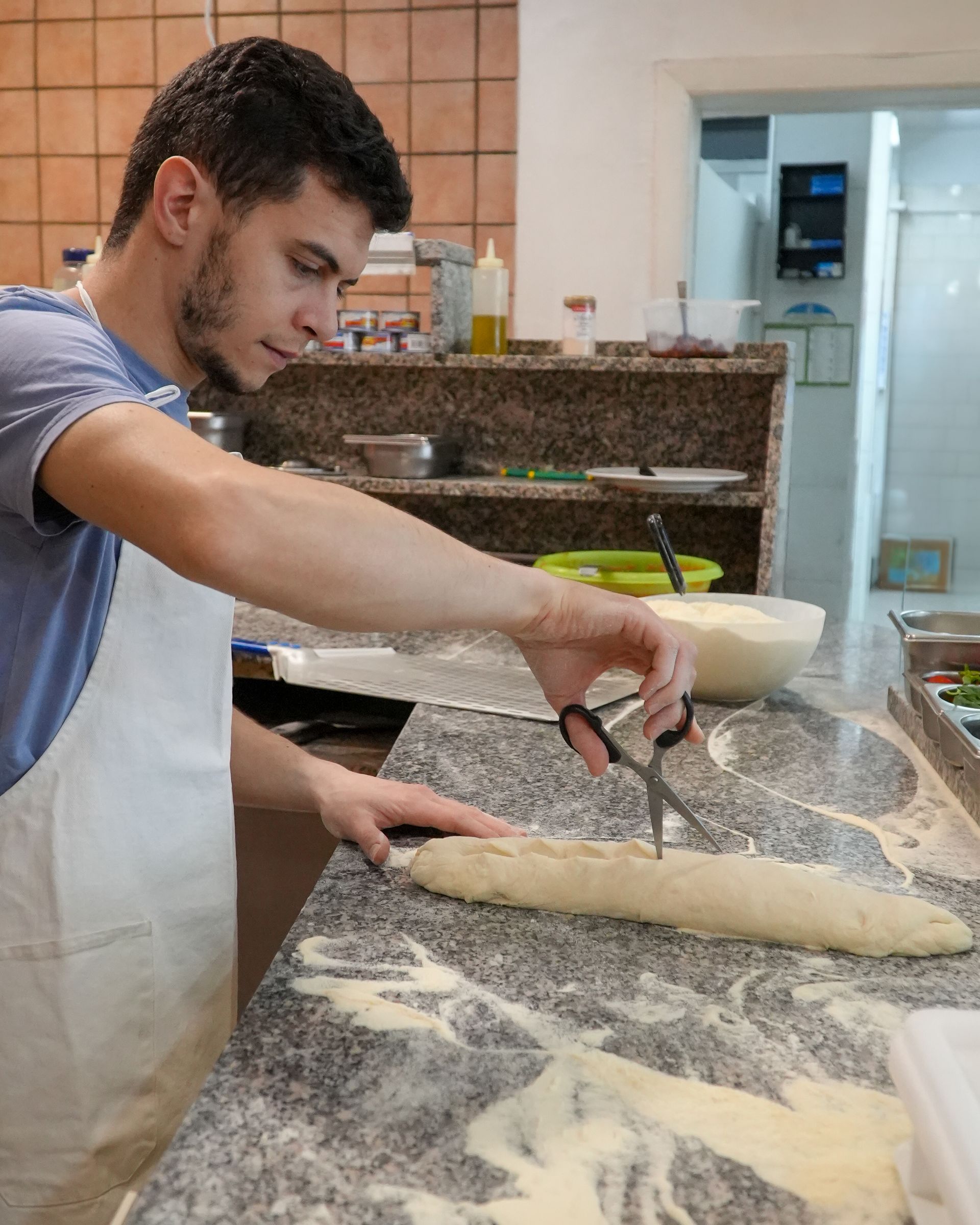 Baker dusting flour and shaping dough on a countertop in a kitchen.