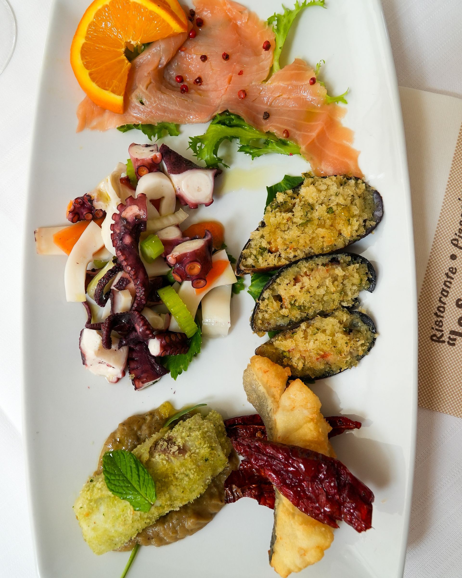 Antipasti assortiti su un piatto bianco: salmone, insalata, bocconcini impanati e salse guarnite con fette d'arancia.