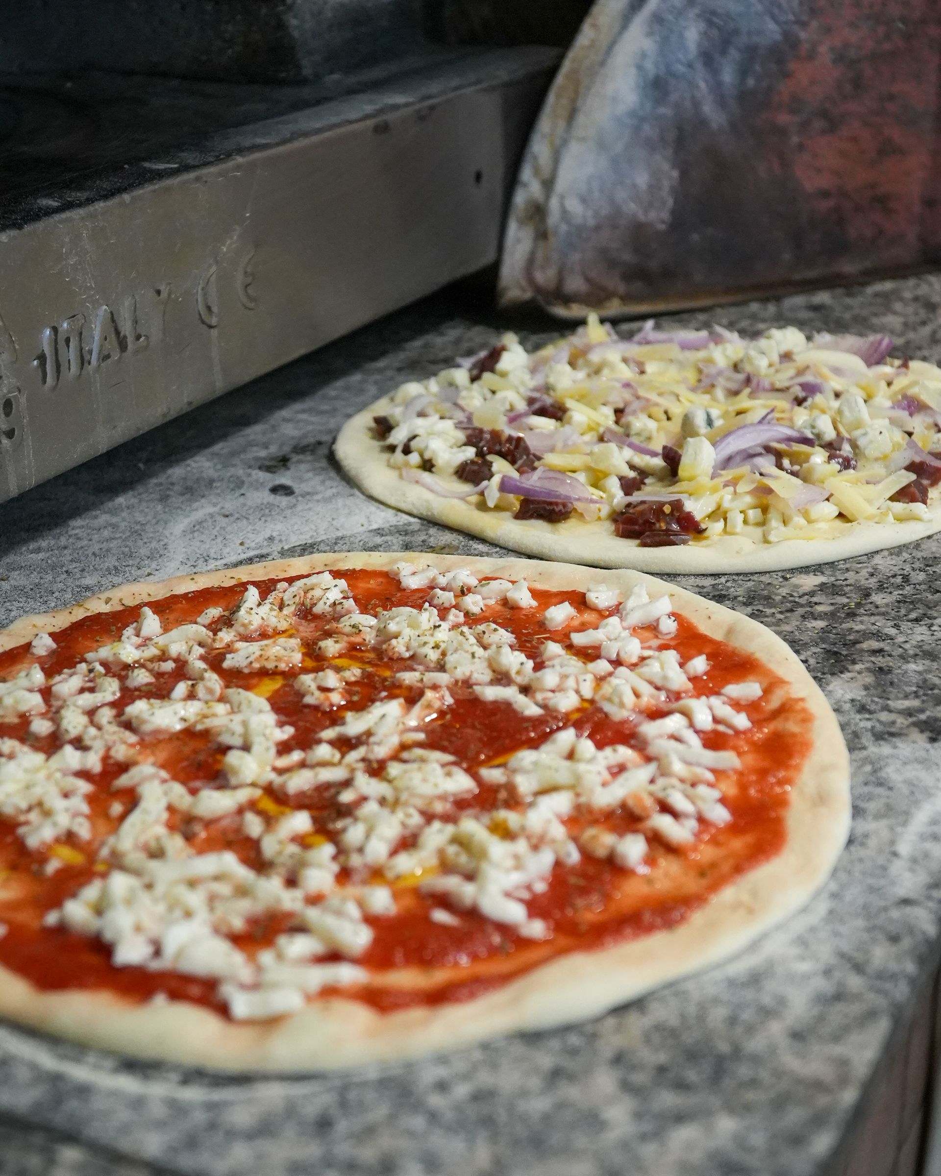 Two uncooked pizzas with tomato sauce, cheese, and toppings on a stone countertop near an oven.