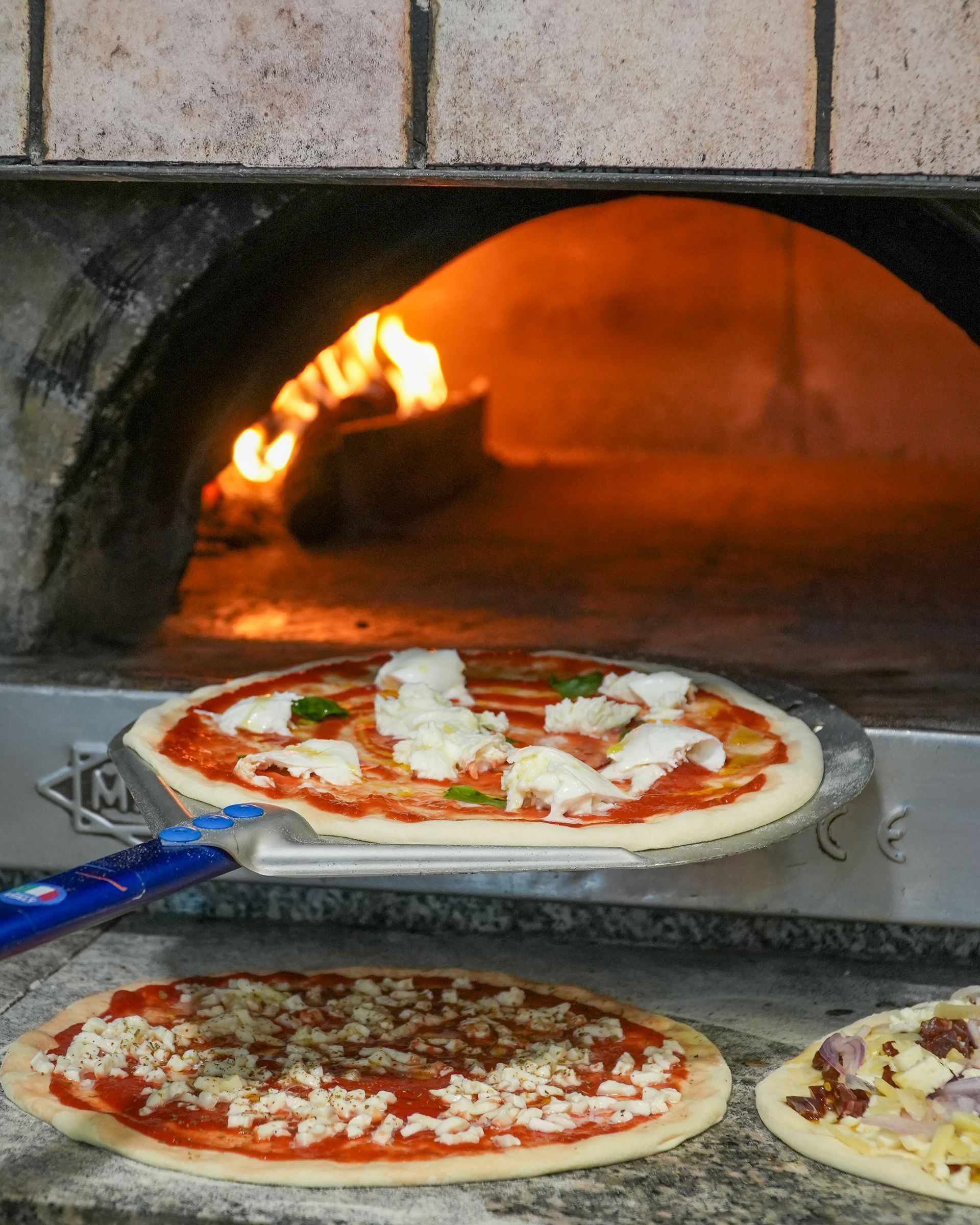 Pizzas baking in a wood-fired oven, with one on a peel in the foreground and flames visible inside.