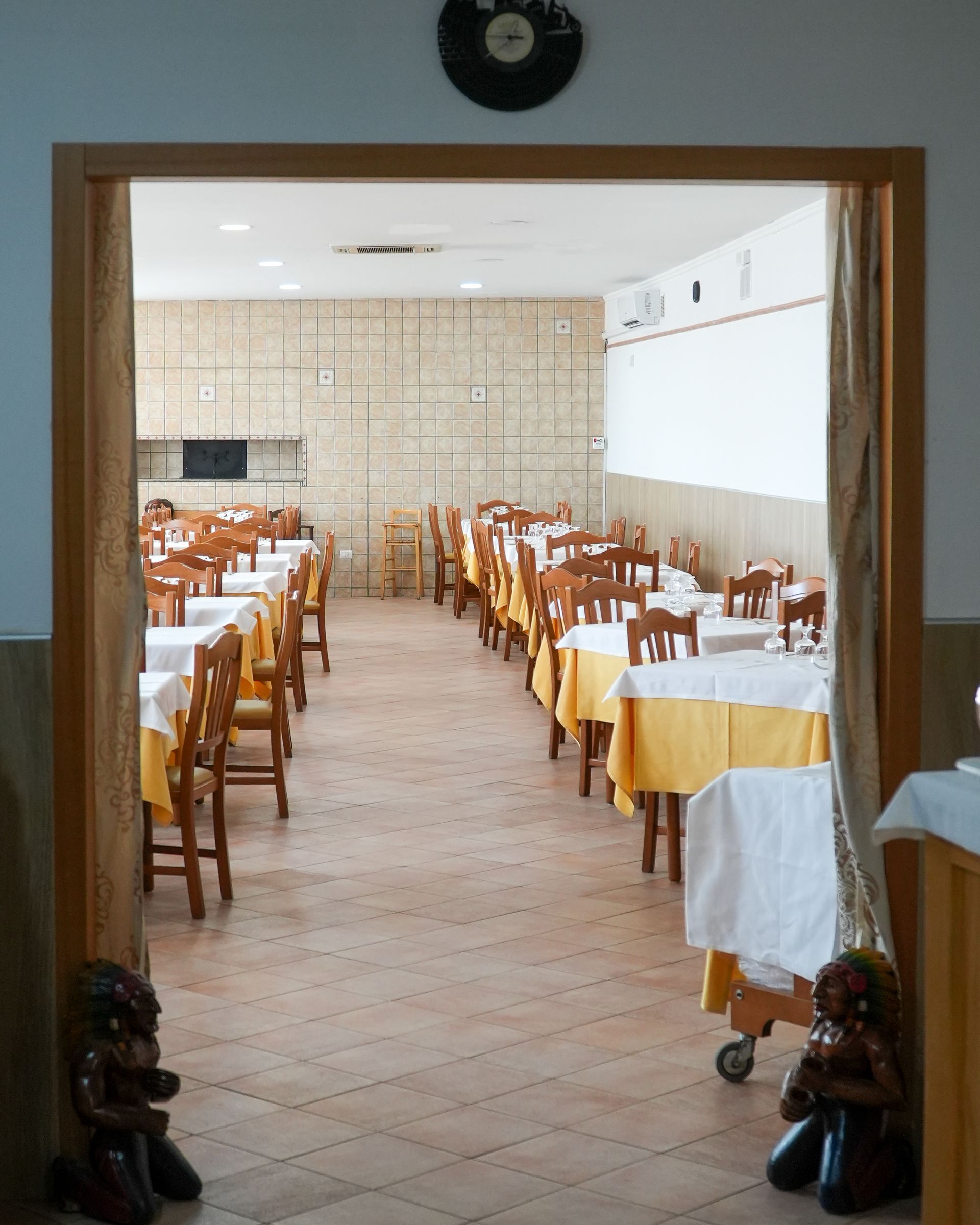 Sala da pranzo vuota di un ristorante con file di sedie e tavoli in legno apparecchiati con tovaglie bianche e gialle.