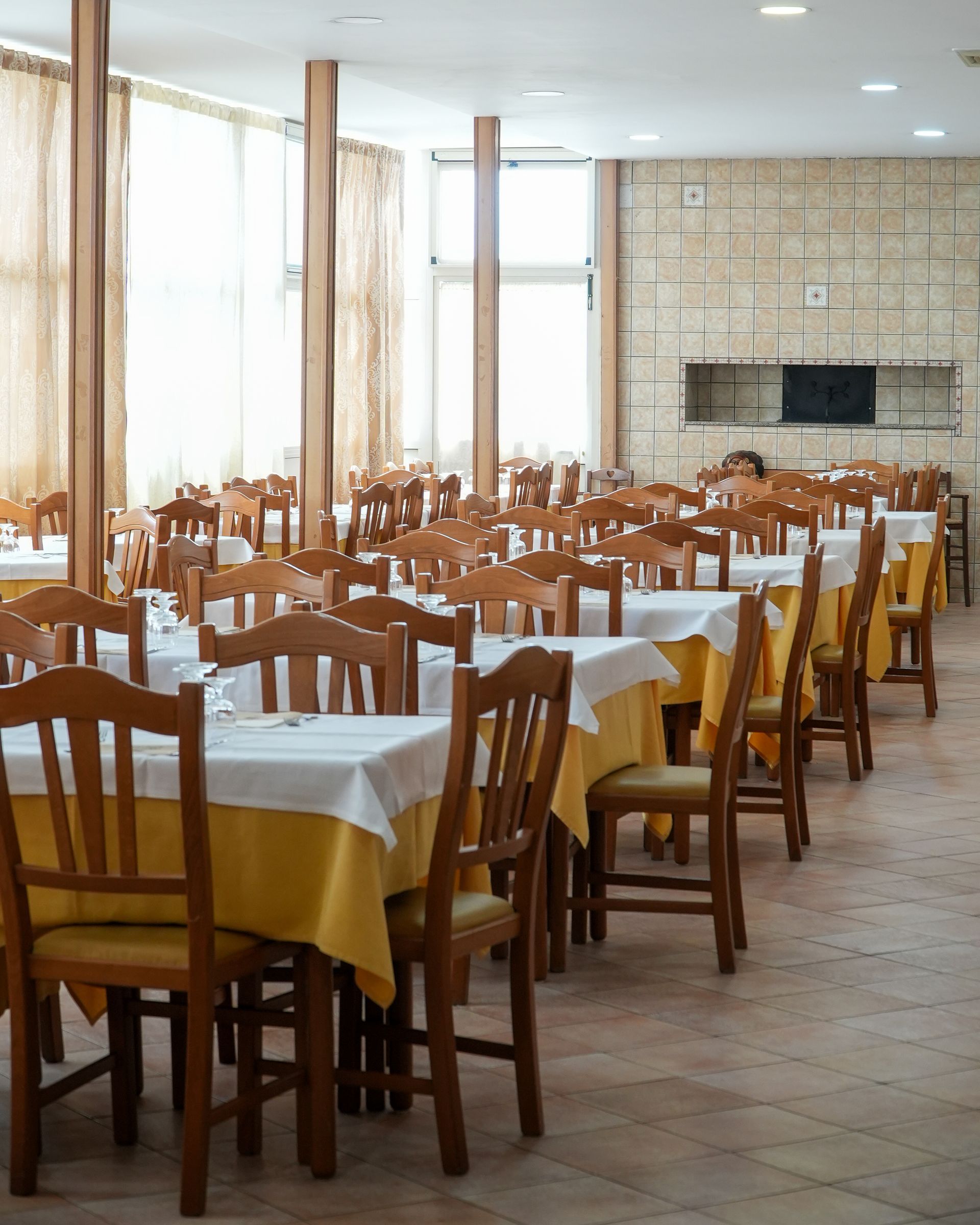 Dining room with rows of wooden chairs and white-tablecloth tables near large windows and a stone fireplace