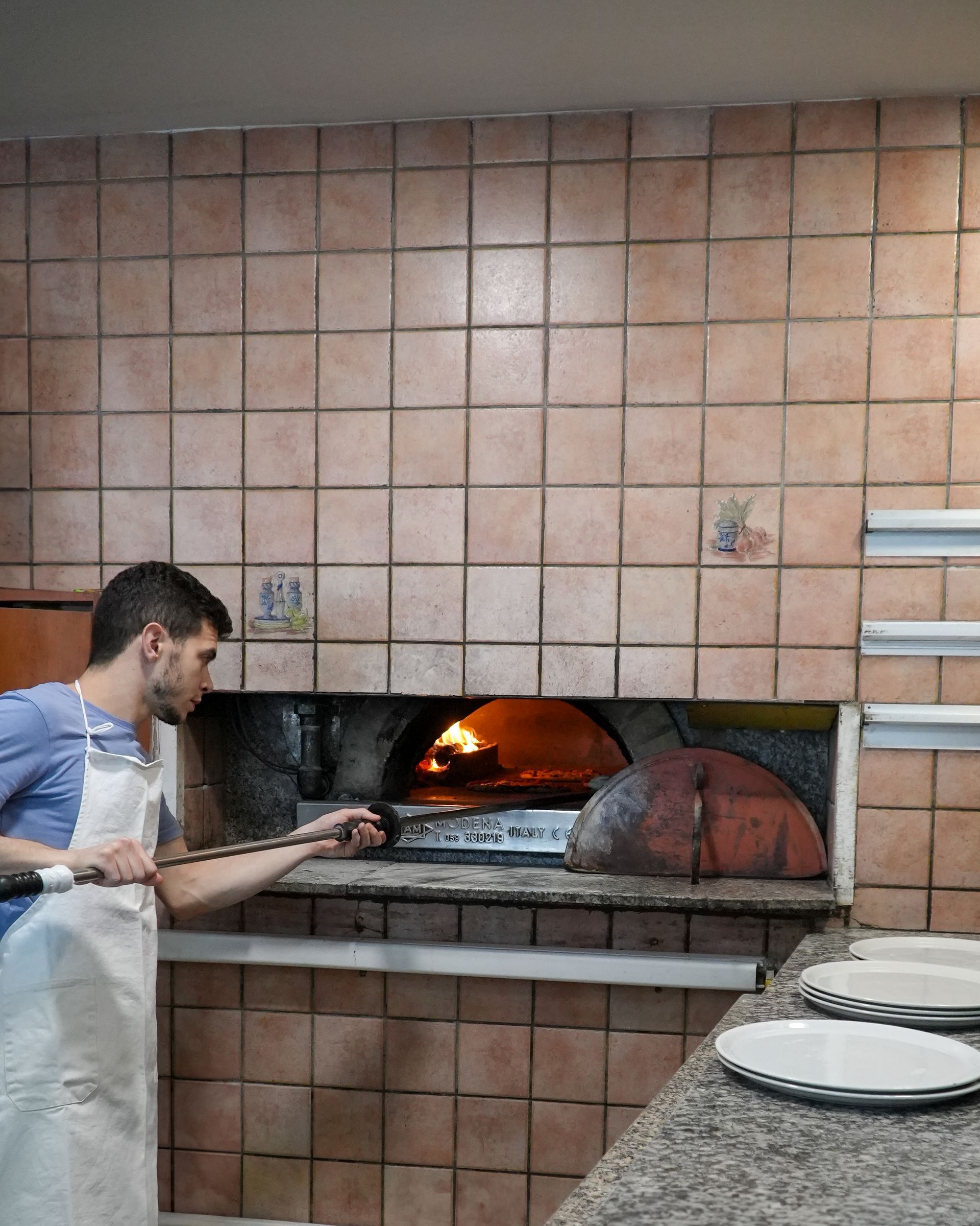 Man tending a wood-fired oven in a tiled bakery, with pizzas on a counter nearby.