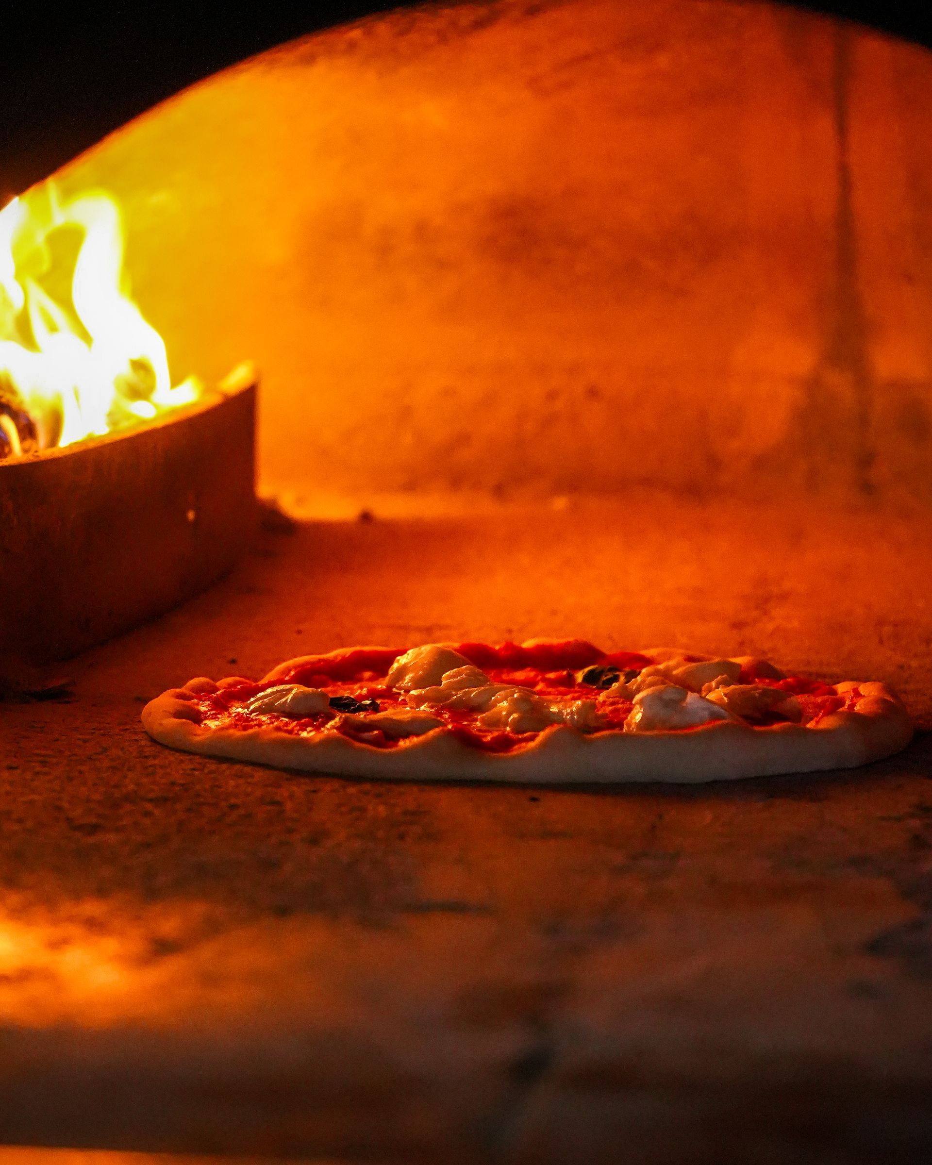 Pizza baking in a wood-fired oven with orange flames and a stone crust.