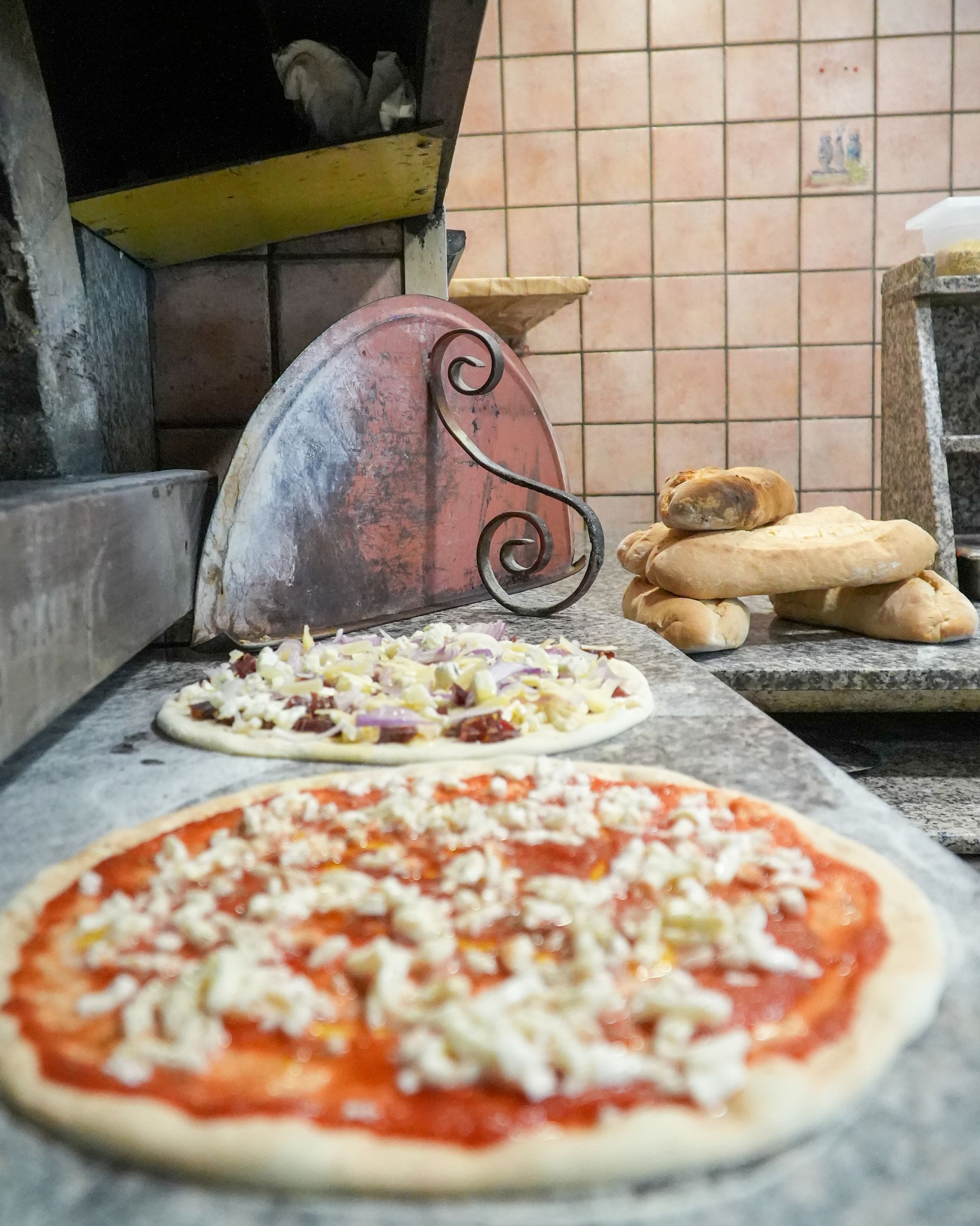 Pizzas preparing in a rustic brick oven kitchen, with dough, toppings, and baked loaves nearby.