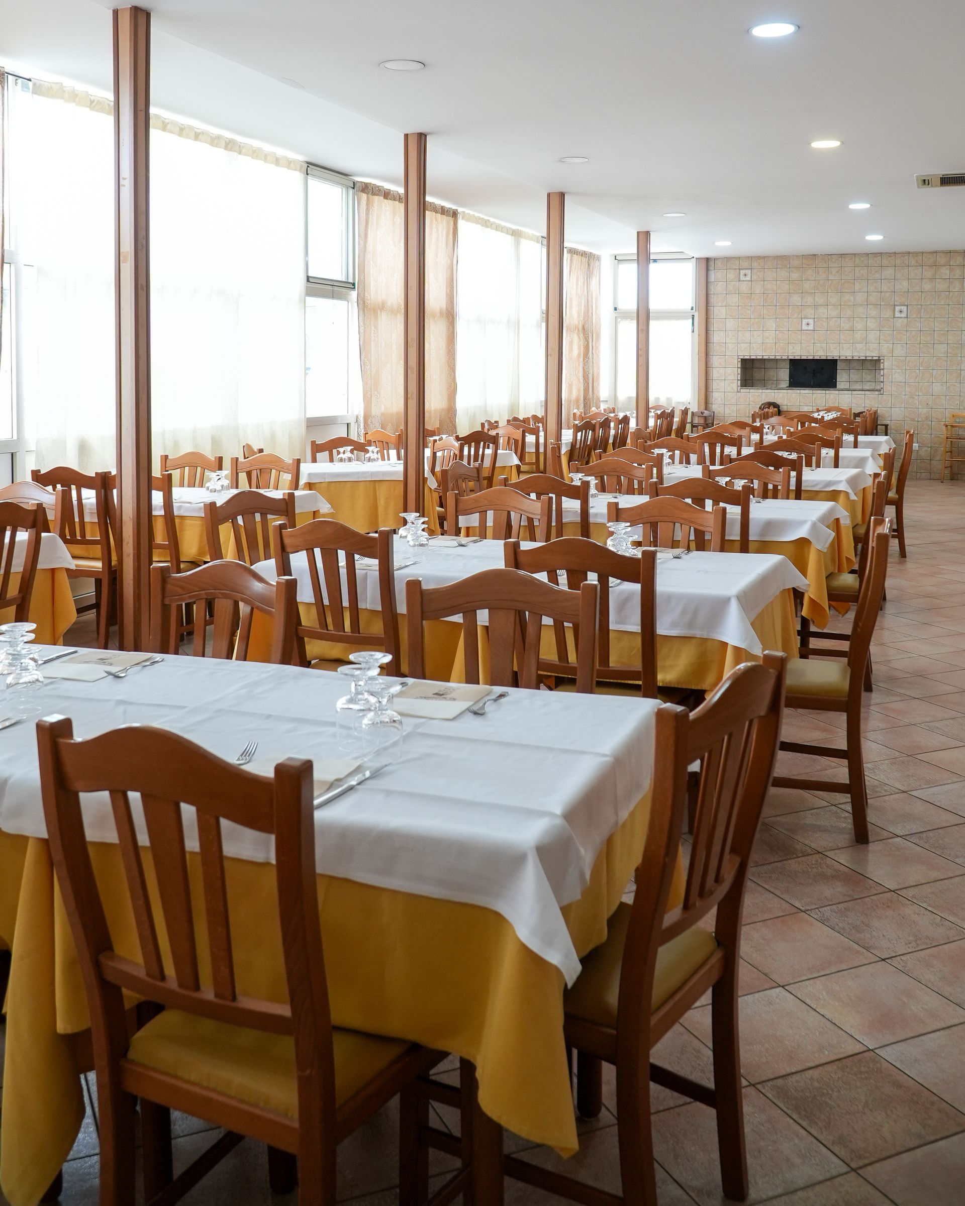 Sala da pranzo vuota con file di sedie e tavoli di legno apparecchiati con tovaglie bianche e gialle.