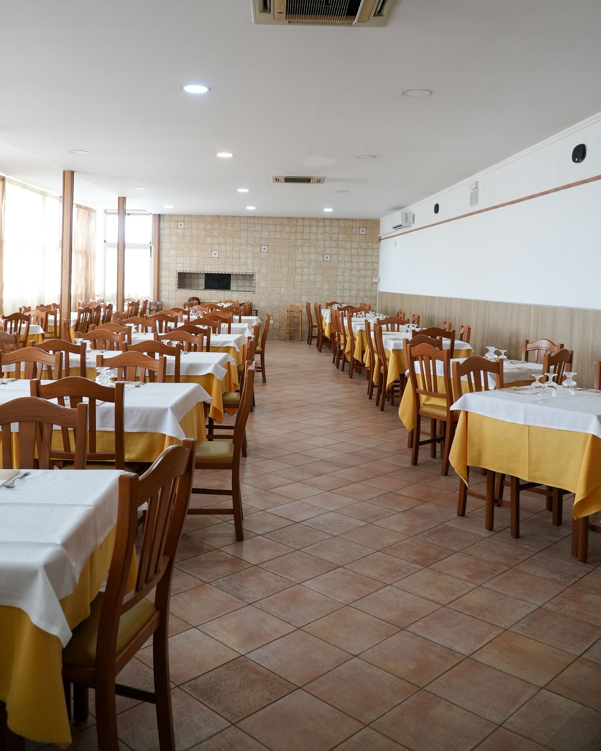 Sala da pranzo vuota di un ristorante con file di sedie di legno, tovaglie bianche e gialle e pavimento piastrellato.