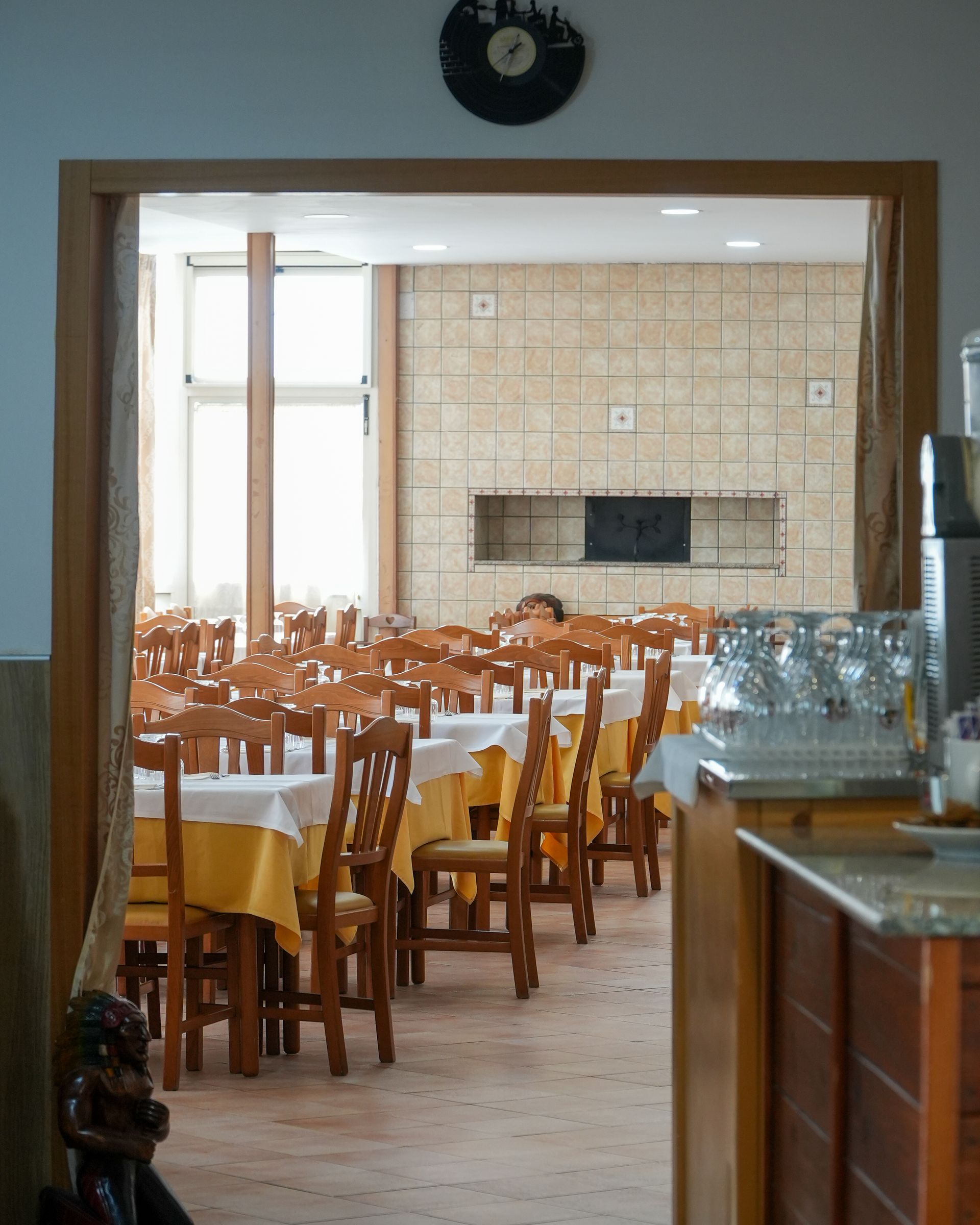 Sala da pranzo vuota con sedie e tavoli in legno, pareti piastrellate e bicchieri su un bancone.