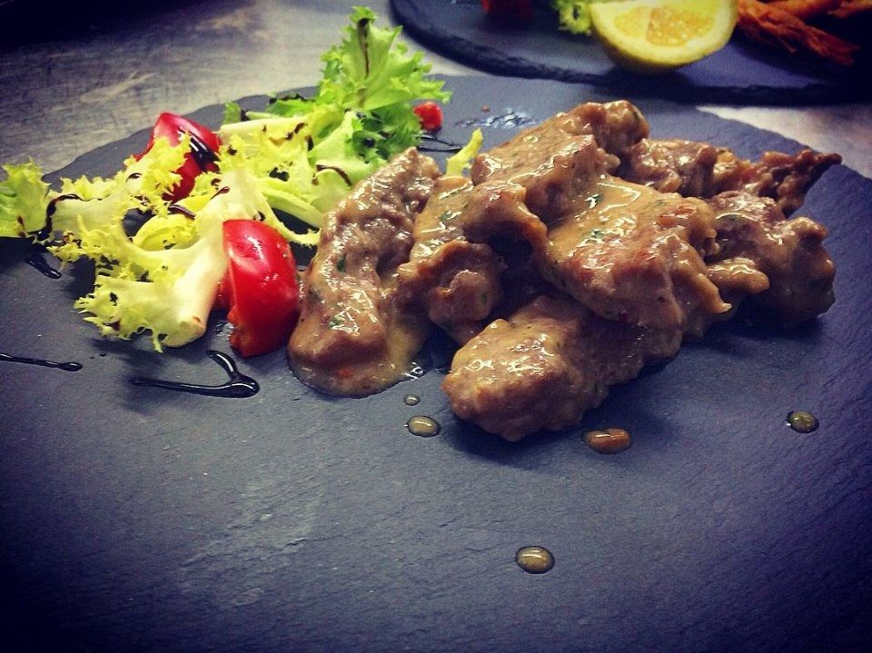 Carne in salsa con una piccola insalata su un piatto scuro, guarnita con pomodorini e spicchio di limone.