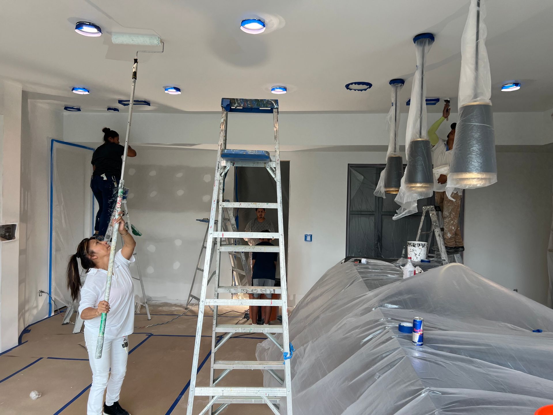 A woman is standing on a ladder in a room with people painting the ceiling.