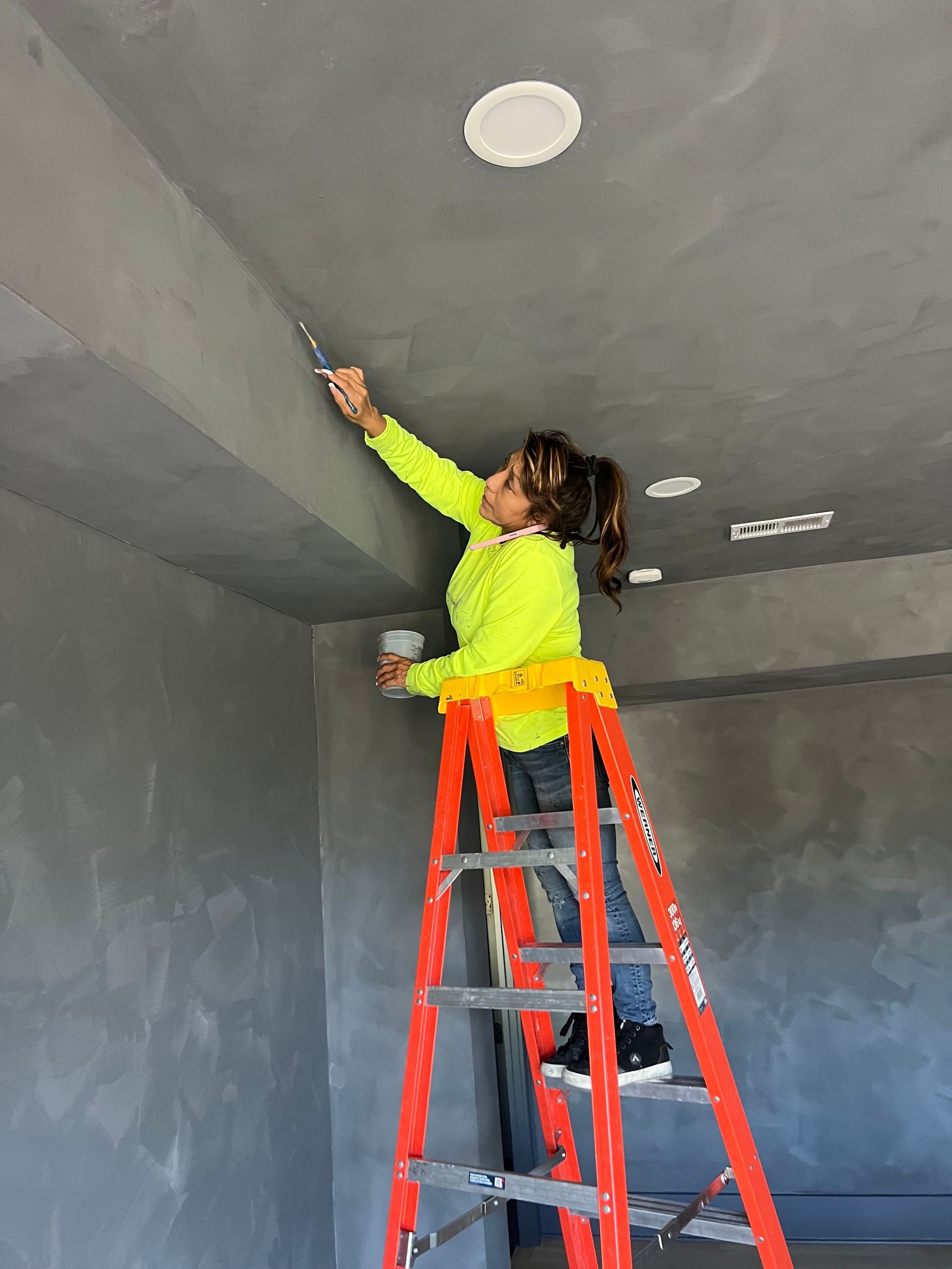 A woman is standing on a ladder painting the ceiling.