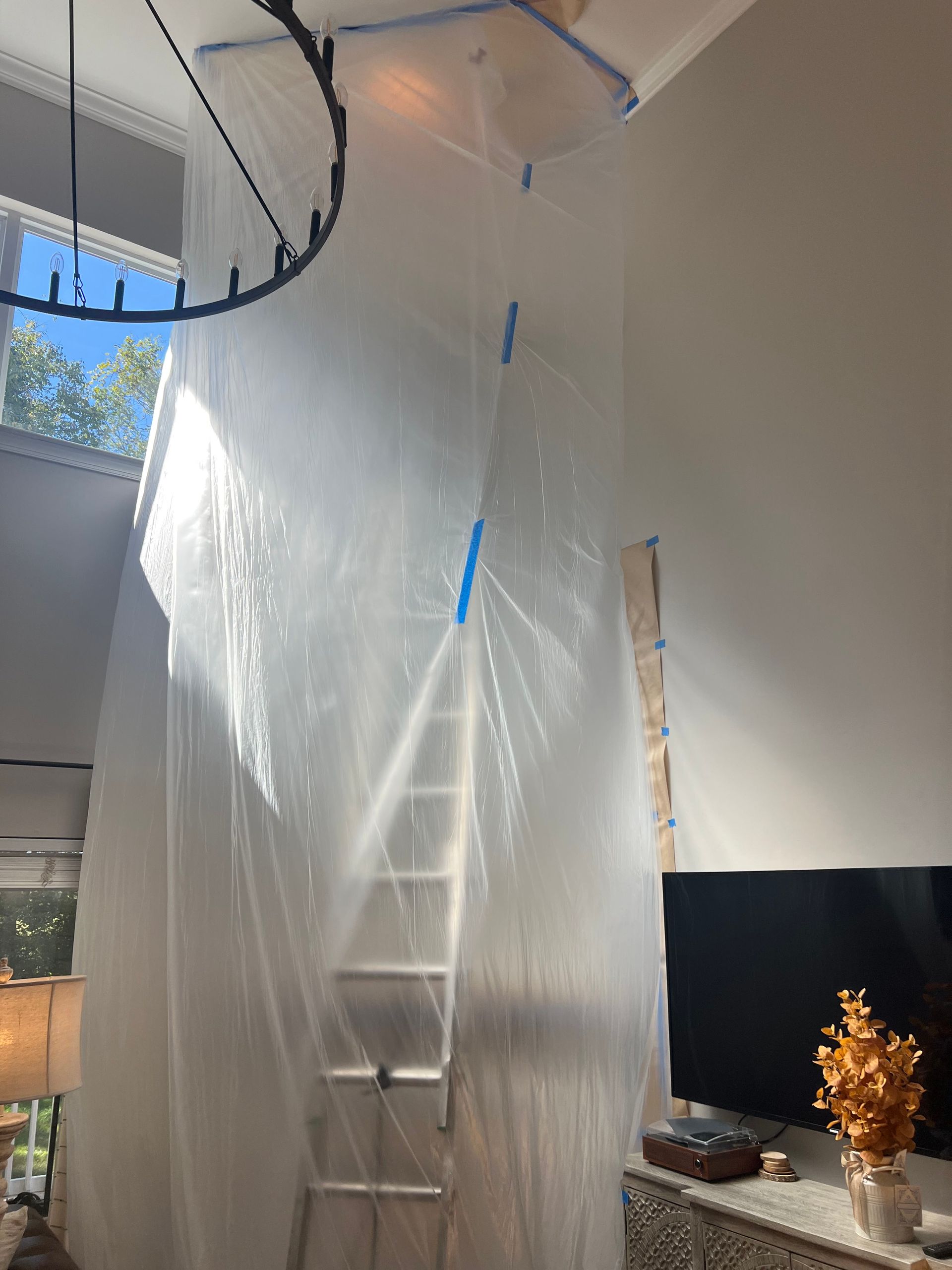 A living room with a ladder covered in plastic and a chandelier hanging from the ceiling.