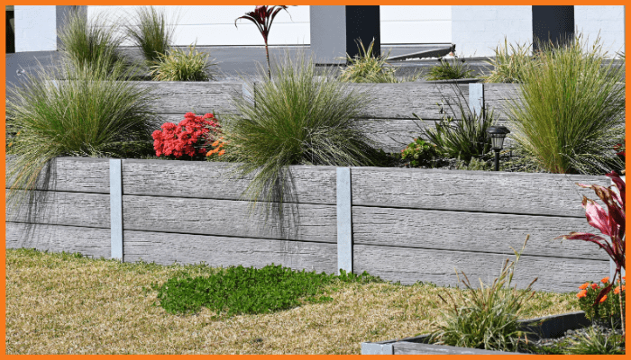 A wooden look concrete retaining wall with plants and flowers in front of a house.