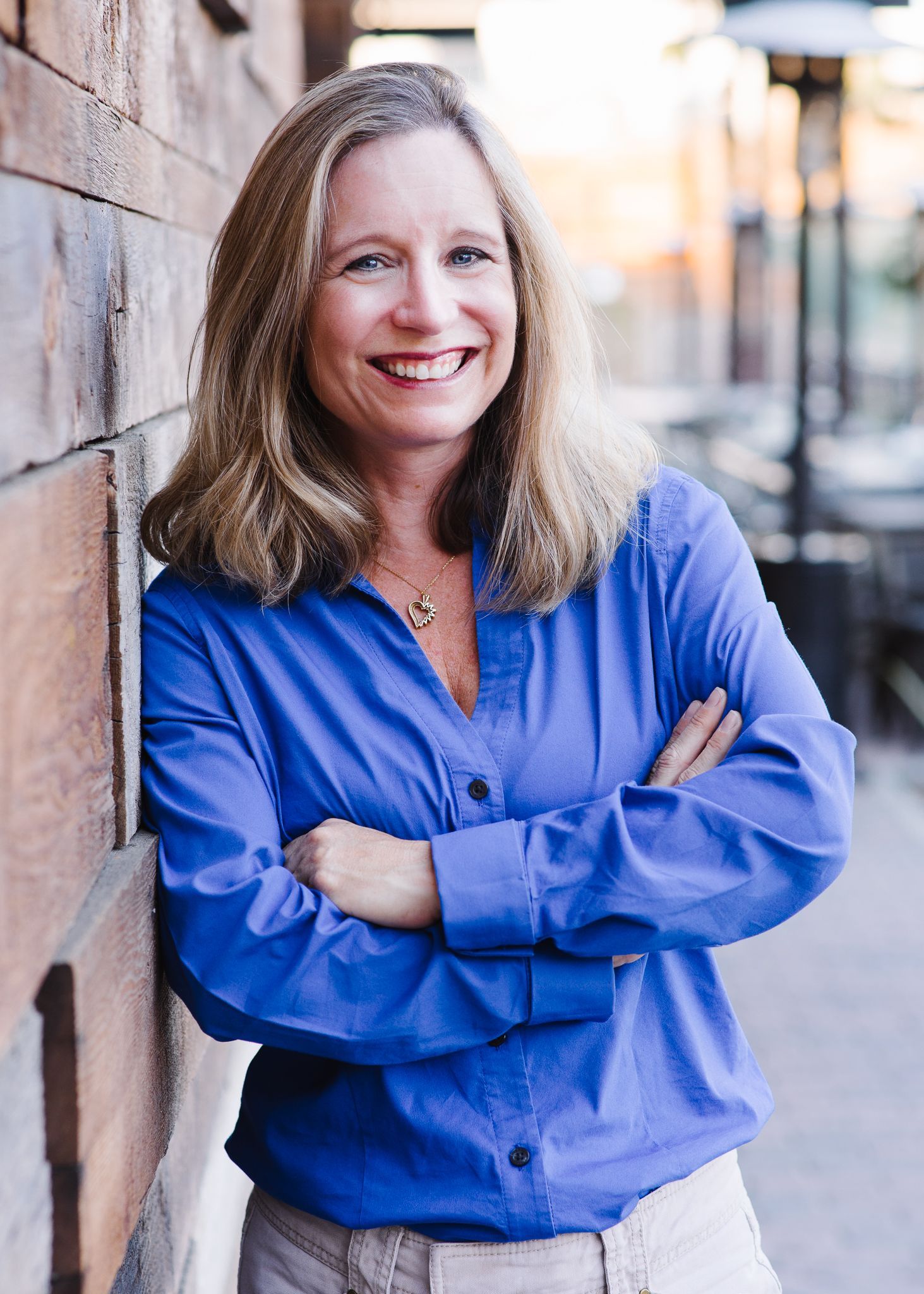 A woman in a blue shirt is leaning against a wall with her arms crossed and smiling.
