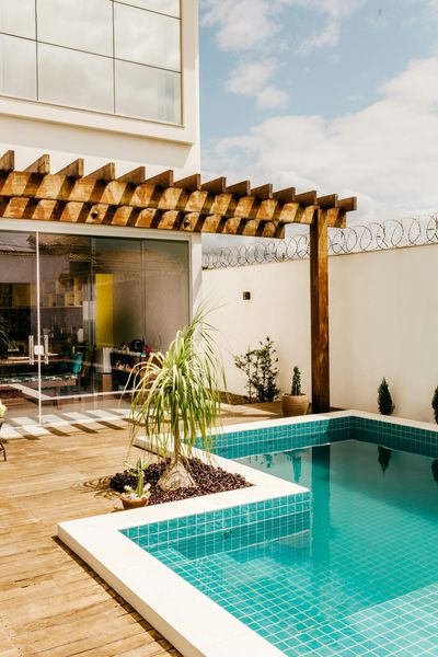 Backyard with pool, wooden pergola, glass wall, plants, and clear blue sky.