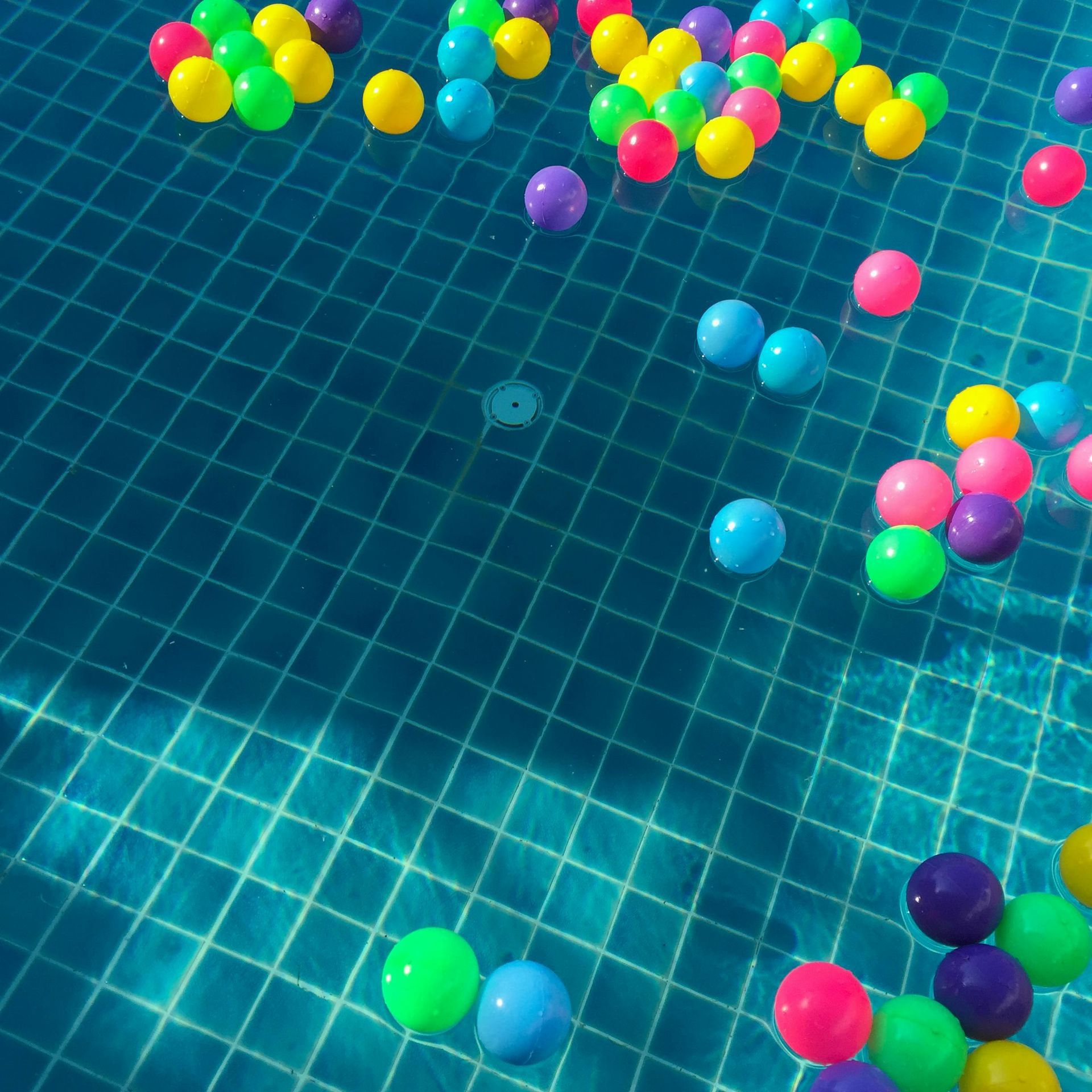 Colorful plastic balls floating in a blue tiled swimming pool, brightly lit by sunlight.