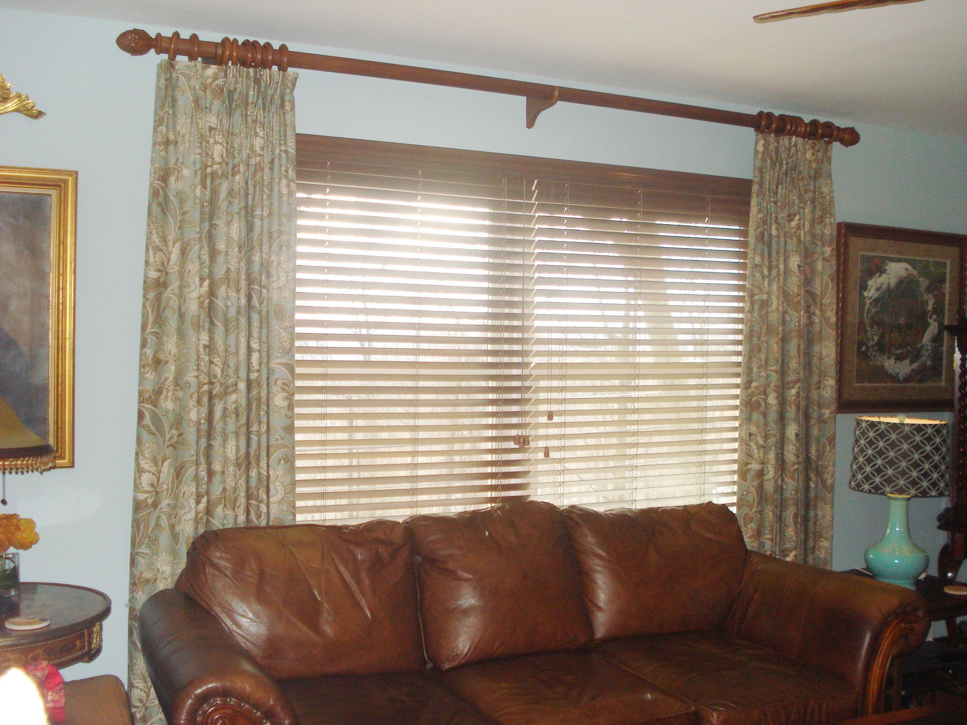 Brown leather sofa in front of a window with wooden blinds and patterned curtains.