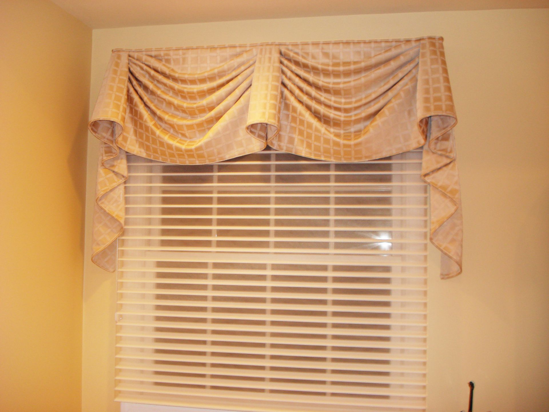 Gold valance draped over a window with horizontal blinds, set against a light yellow wall.