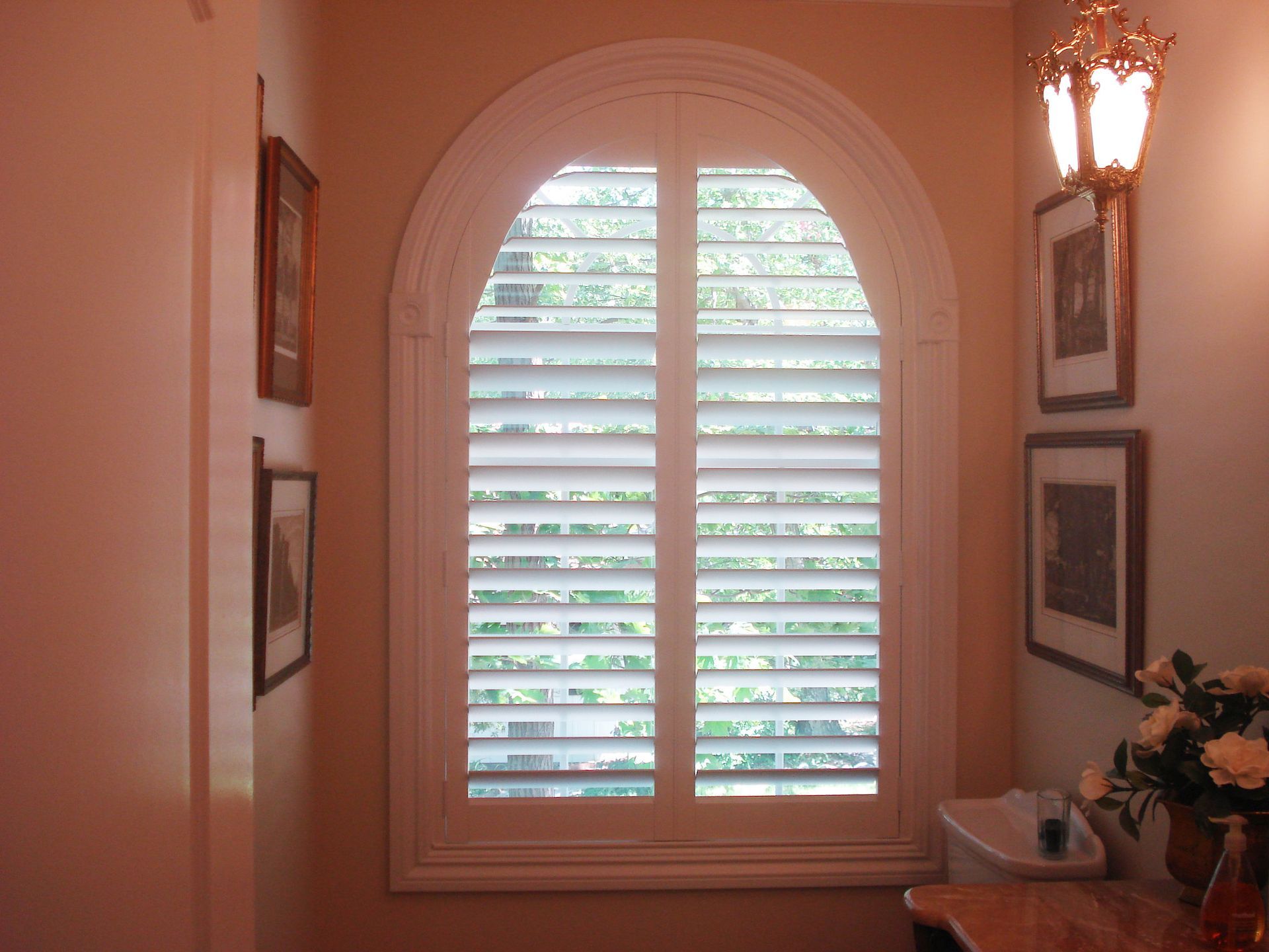 Arched window with white shutters, framed by a decorative molding. Small wall art and a lamp are nearby.