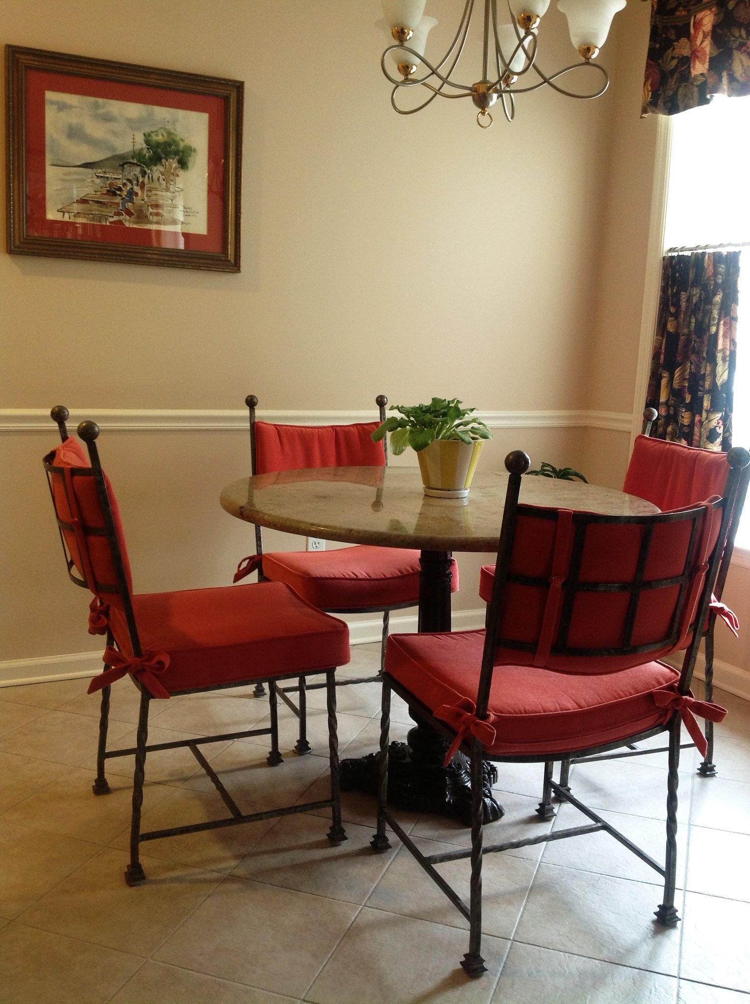 A dining table with four red chairs, beneath a chandelier and painting.