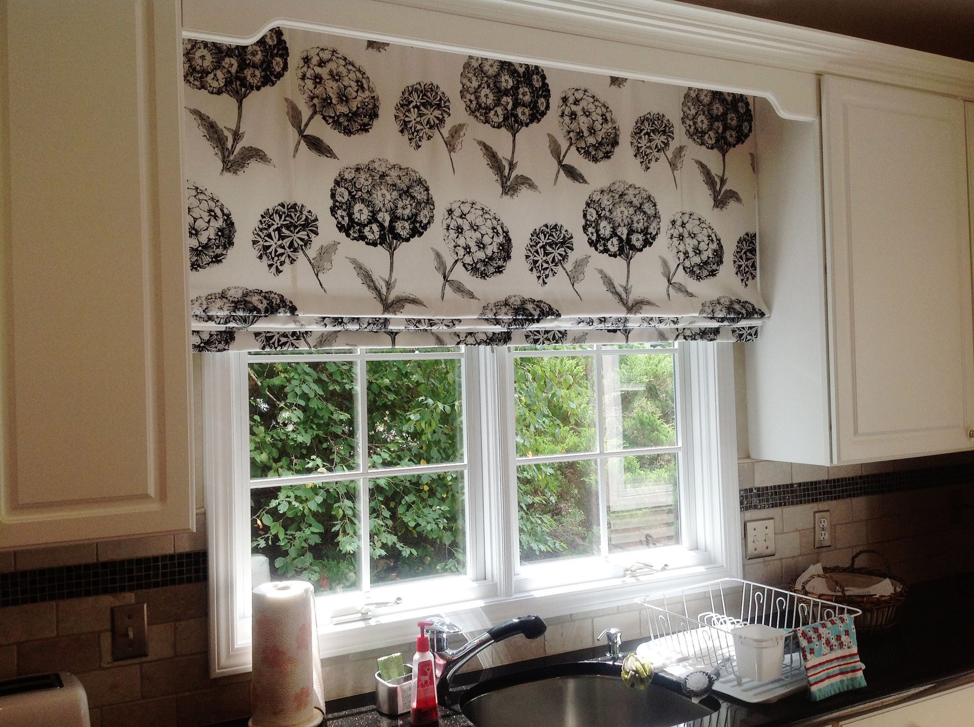 Black and white floral patterned window shade in a kitchen with cabinets and sink.