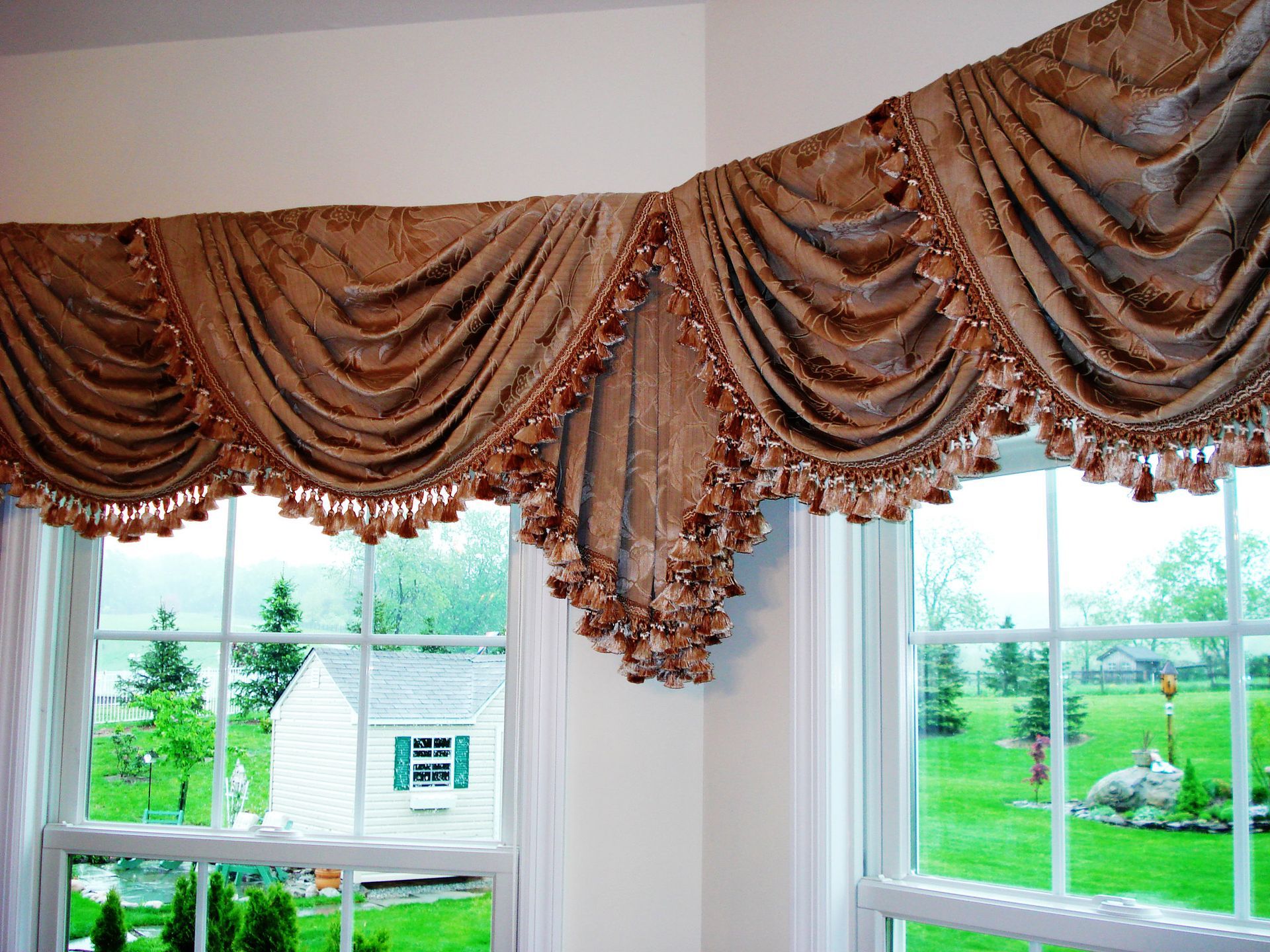 Beige, ornate valance with tassel trim above two windows overlooking a green yard.