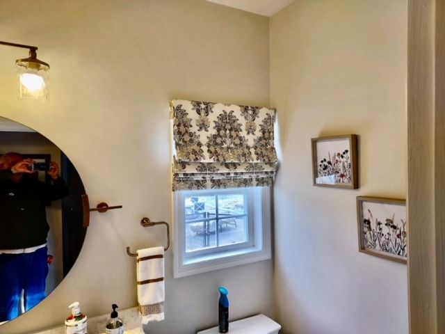 Small bathroom with round mirror, toile window shade, framed art, and towel rack.