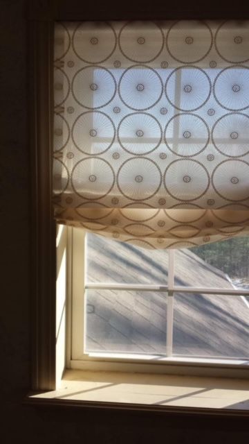 Window with cream-colored Roman shade, featuring a circle pattern. Sunlight streams through the glass.