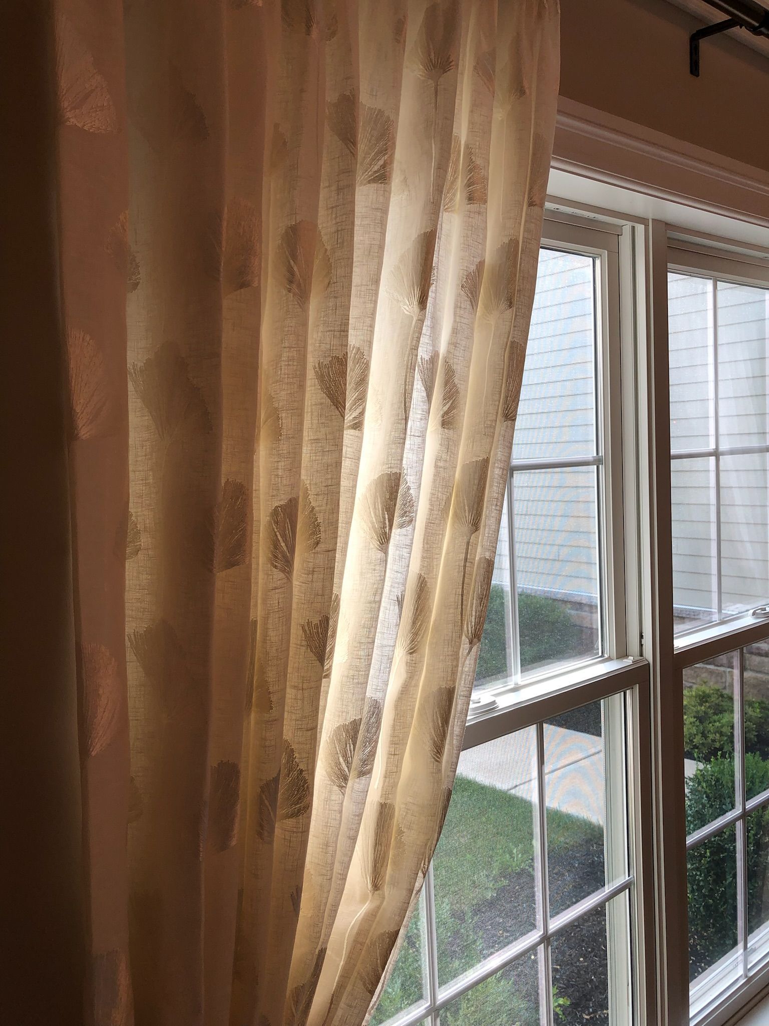Sheer cream-colored curtain with patterned design hangs in front of a window overlooking a green outdoor area.