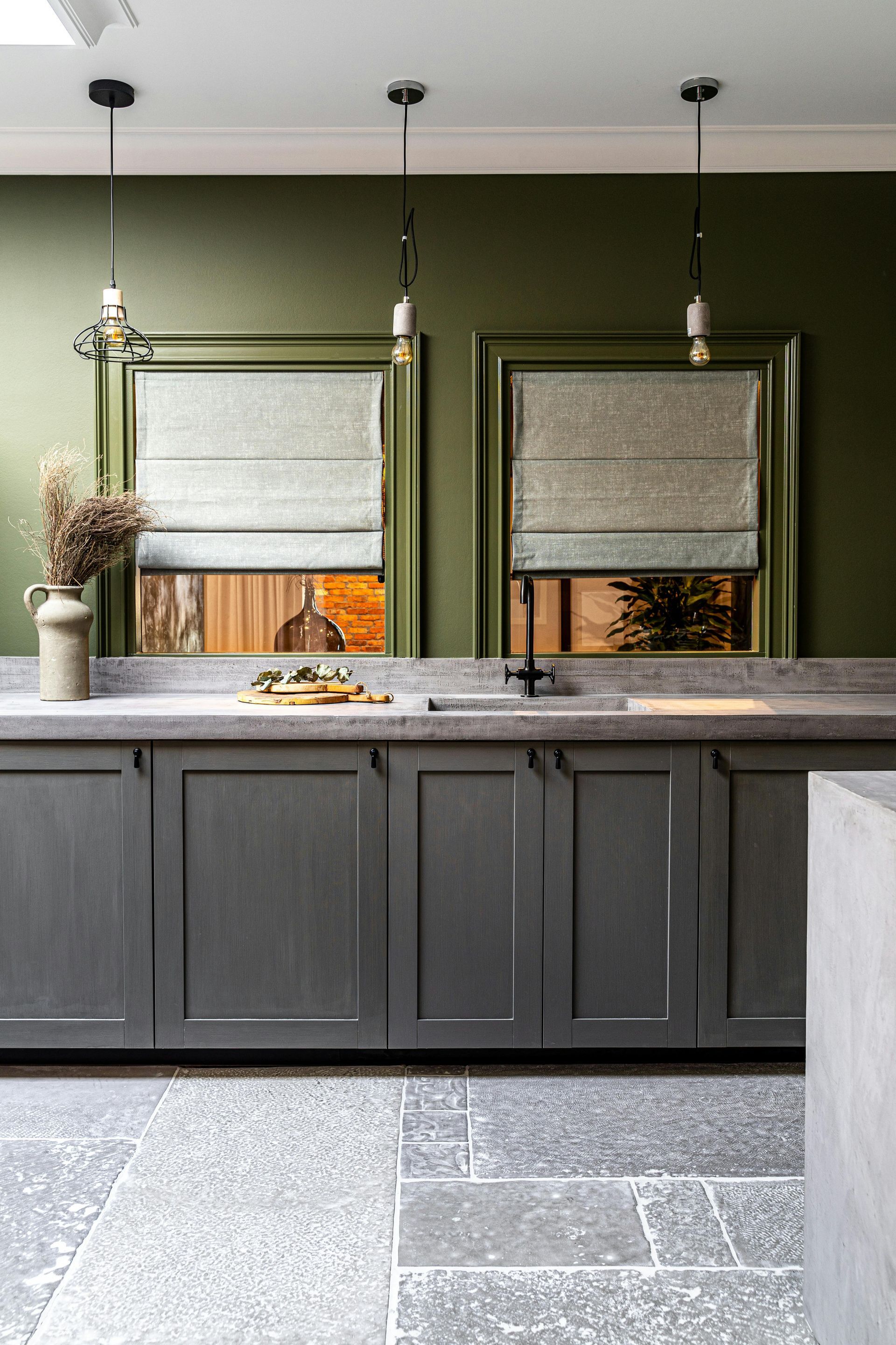 Green-walled kitchen with gray cabinets, countertops, and stone floor. Two windows with patterned blinds.