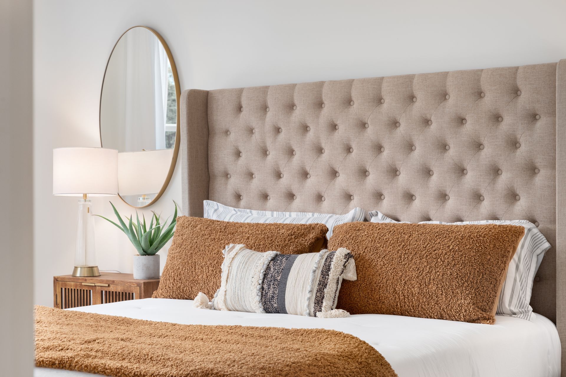 Bedroom with tan tufted headboard, pillows, and gold-framed mirror.