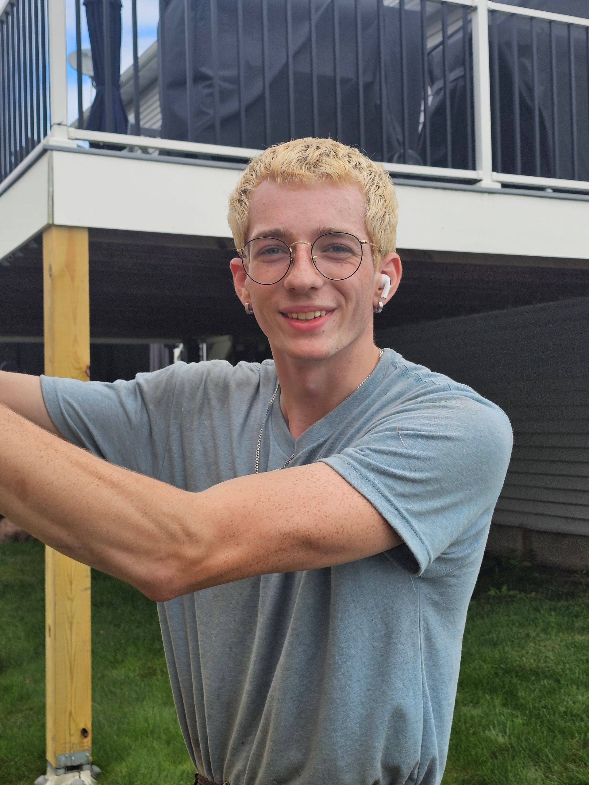 Young man with blonde hair and glasses smiles outdoors, wearing gray shirt.