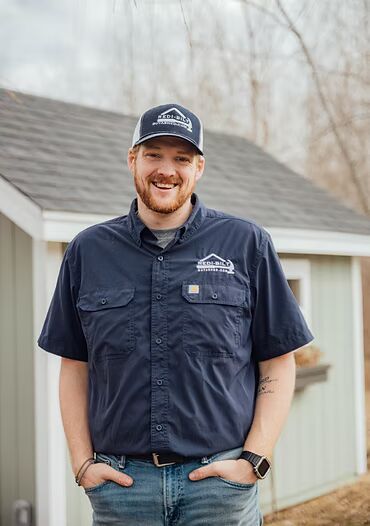 A smiling man with a beard wearing a navy blue work shirt and hat stands in front of a small building.