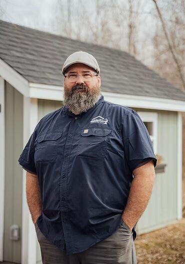 A bearded man in a blue shirt and cap stands in front of a small shed, hands in pockets.
