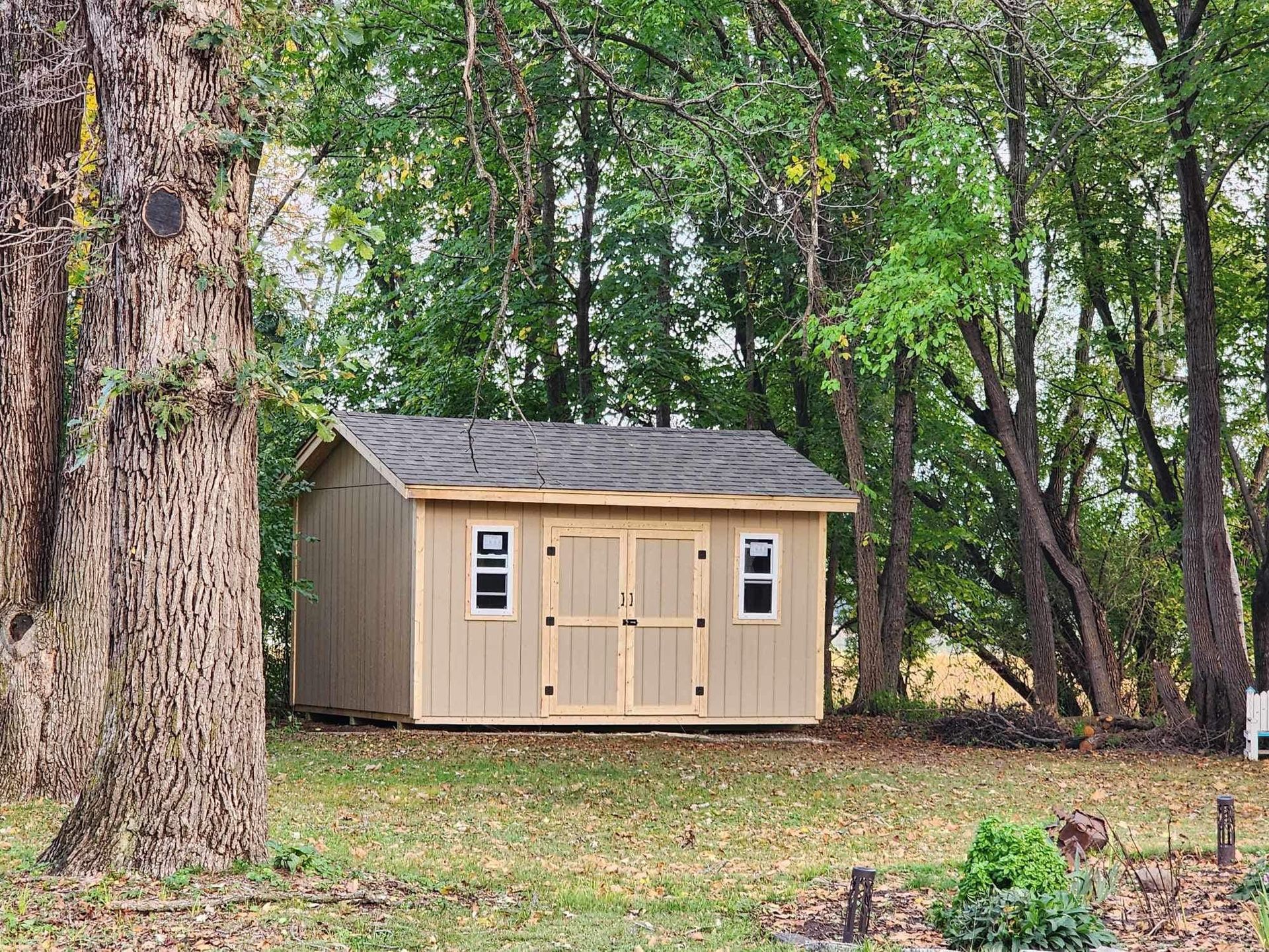 Tan shed with gray roof in a grassy yard surrounded by trees.