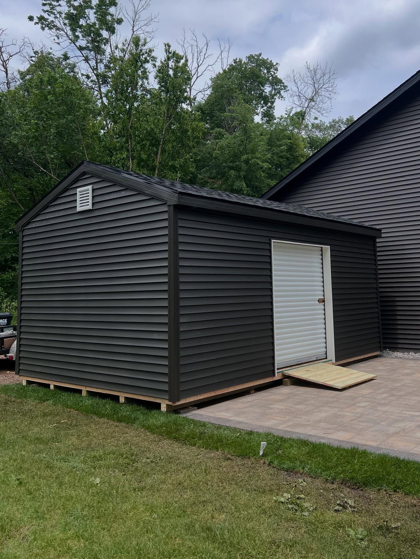 Dark gray shed with a white door and ramp on a paved patio.